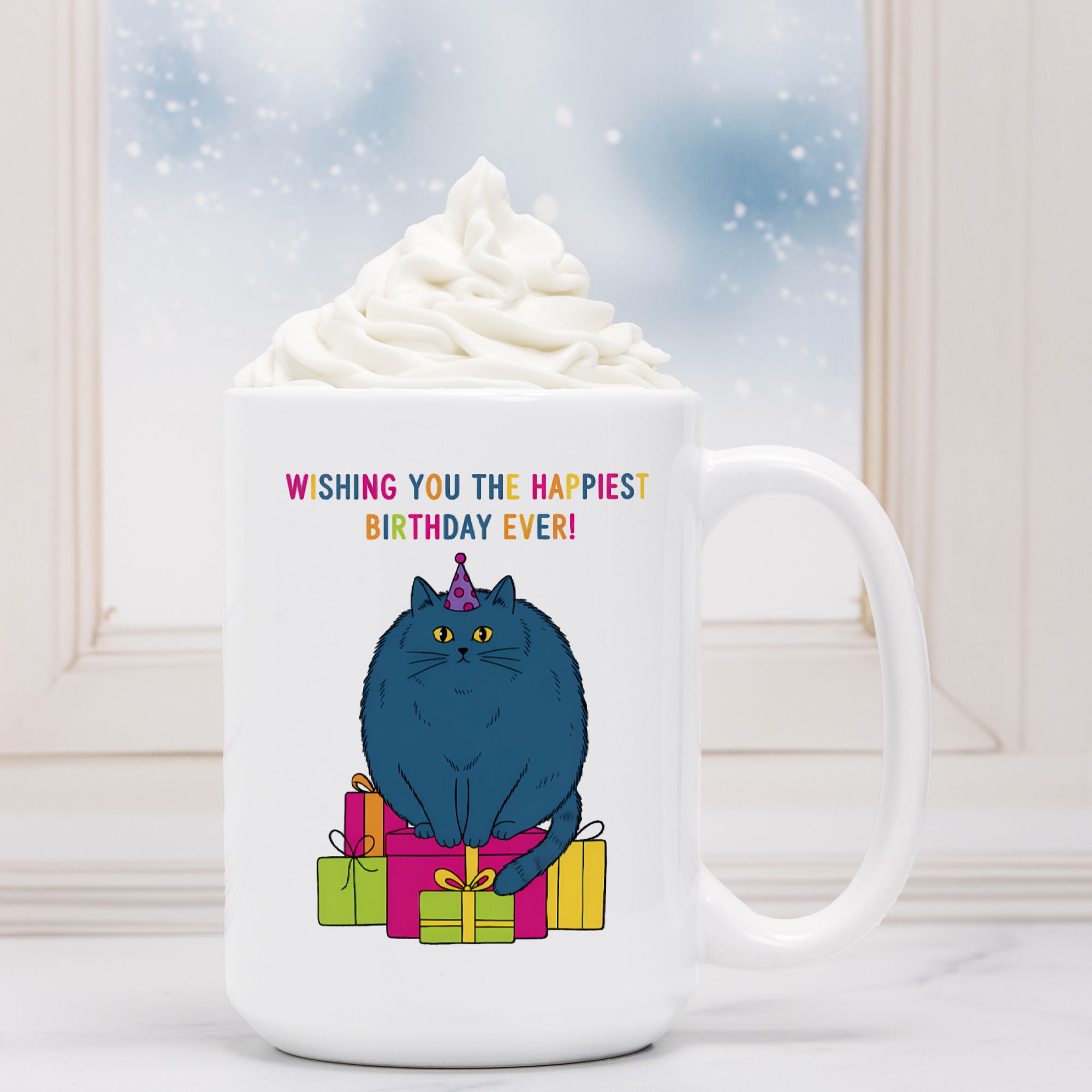 White mug with a blue cat illustration and birthday text, surrounded by whipped cream and hot chocolate.