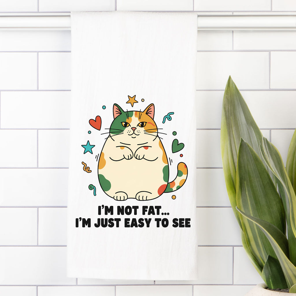 Tea towel with Cartoon cat and text ' I'm Not Fat... I'm Just Easy To See' on a white tiled wall with a plant.