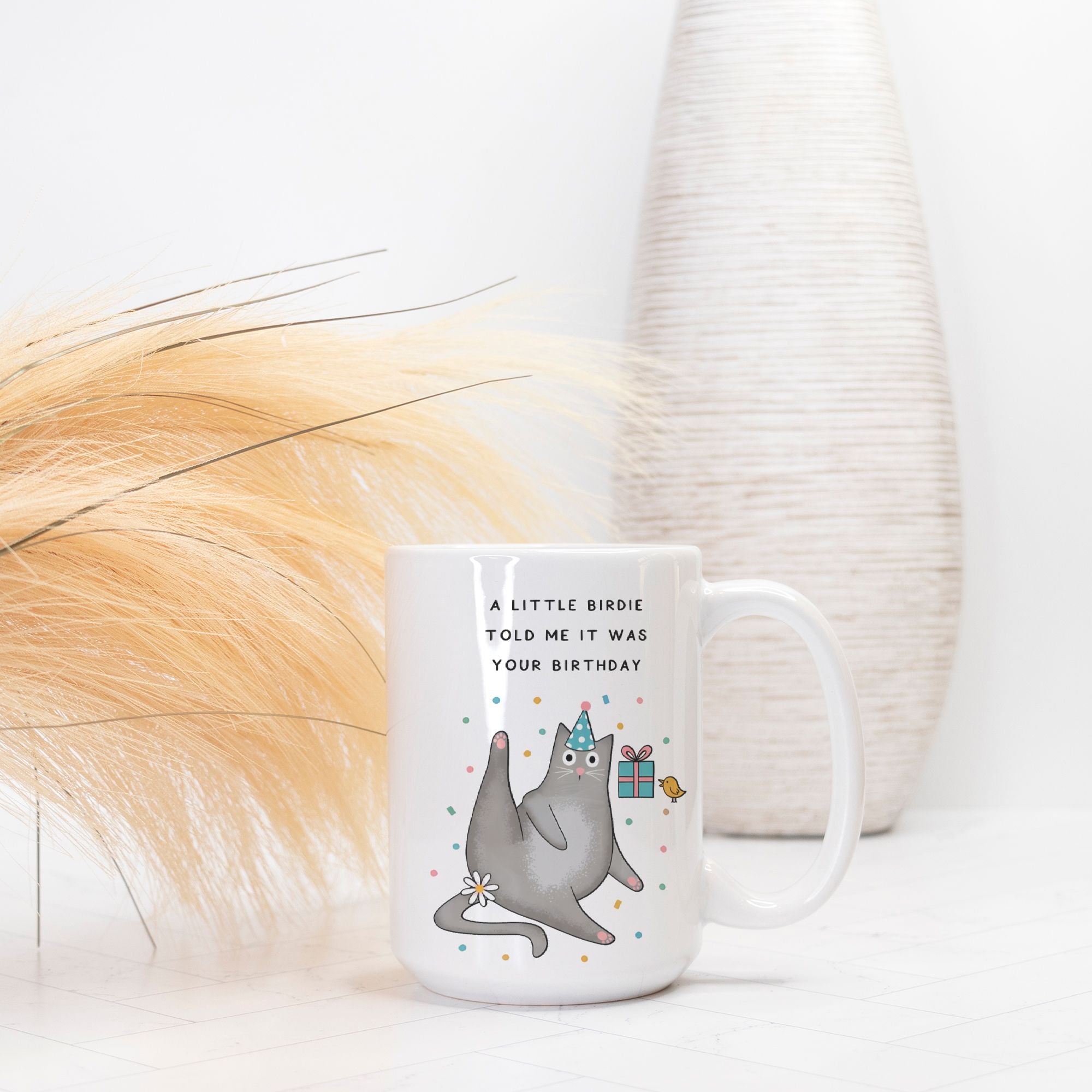 White mug with a cartoon cat wearing a party hat and text 'A little birdie told me it was your birthday' on a white background.
