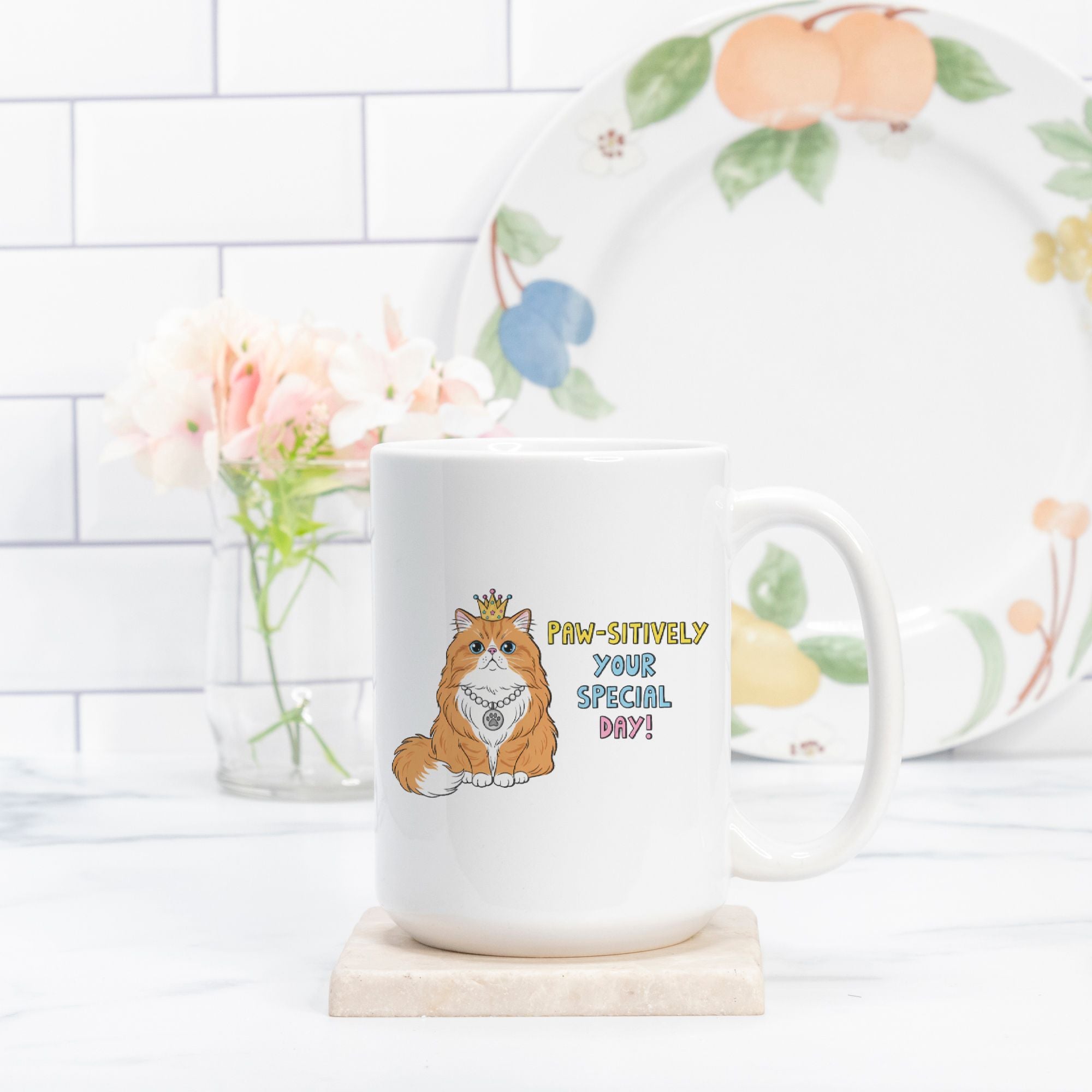 White mug with a cat design and 'Paw-Sitively Your Special Day!' text on a white surface with a floral plate in the background.