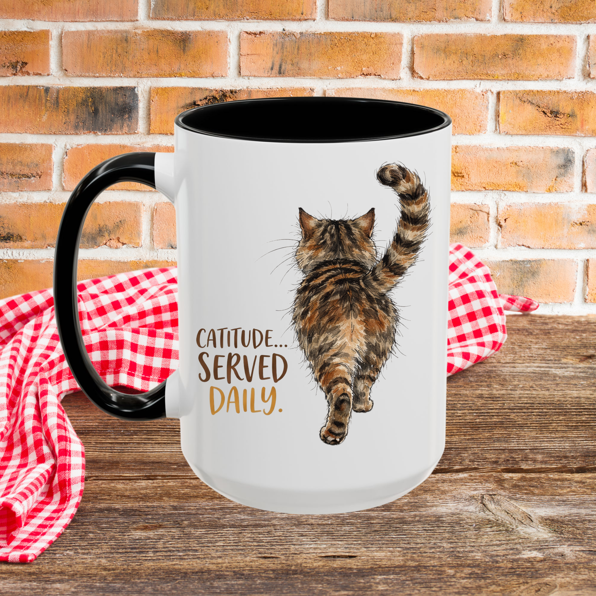 White mug with a cat illustration and 'Catitude... Served Daily' text on a wooden picnic table with a brick wall background.