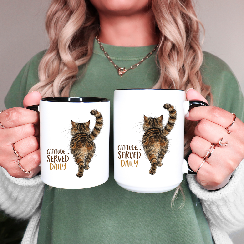 Person holding two black accent mugs with cat illustrations and text 'Catitude Served Daily'.