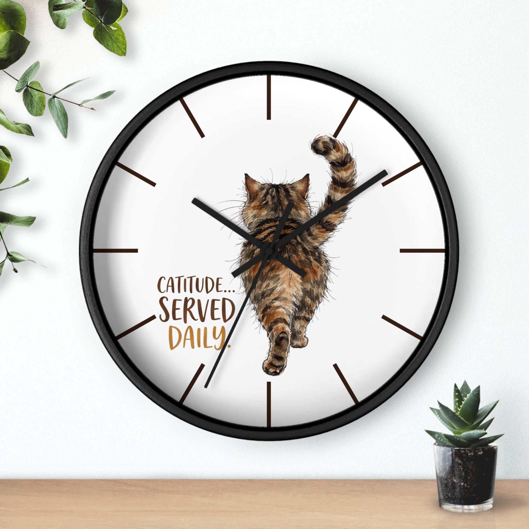 Round black wall clock with a cat design and 'Catitude Served Daily' text, placed on a surface with plants.