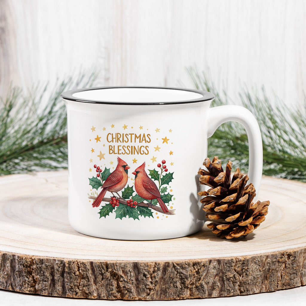 White mug with Christmas cardinals design on a wooden surface with a pine cone and evergreen branches.