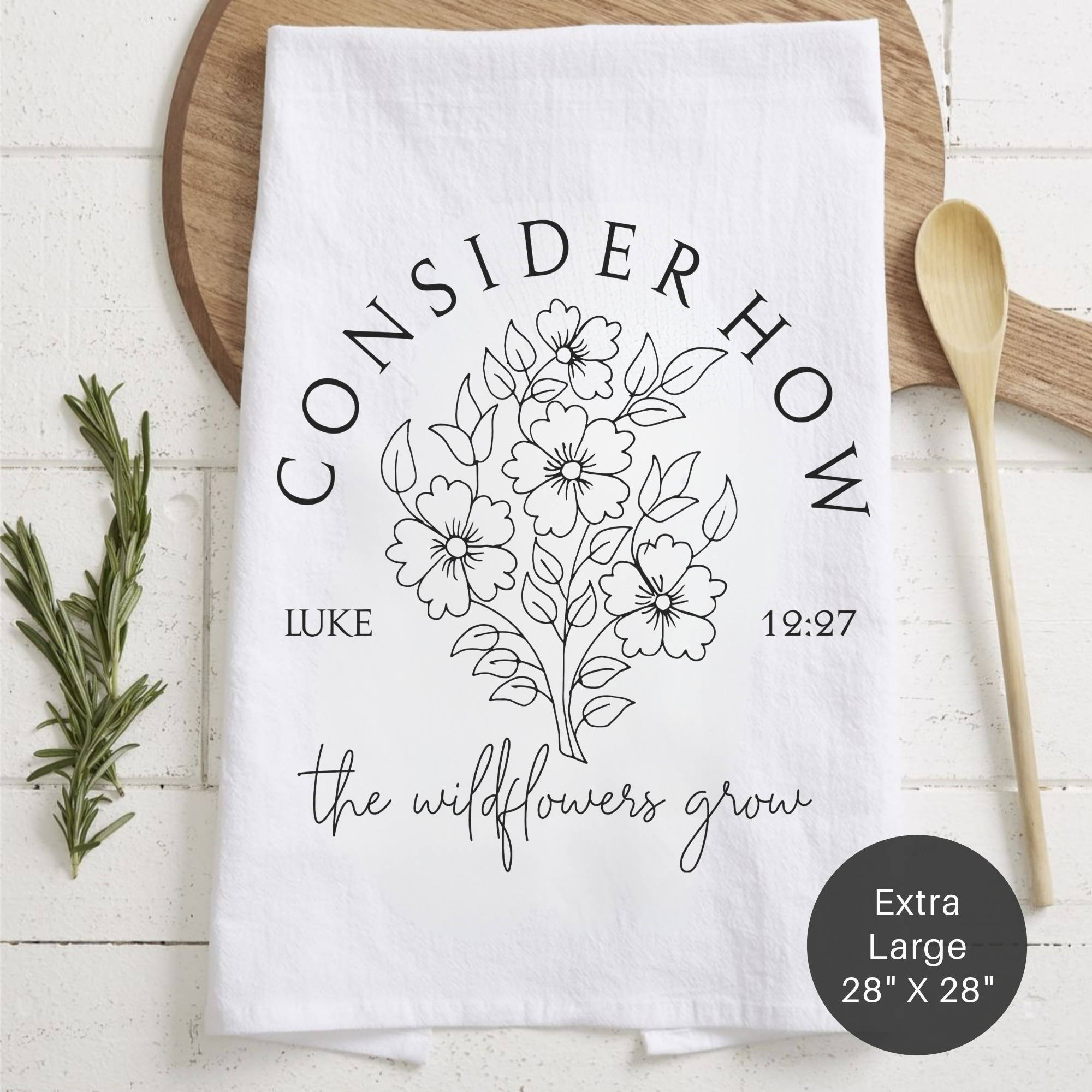 White tea towel with floral design and text ' Consider How The Wildflowers Grow' on a wooden surface