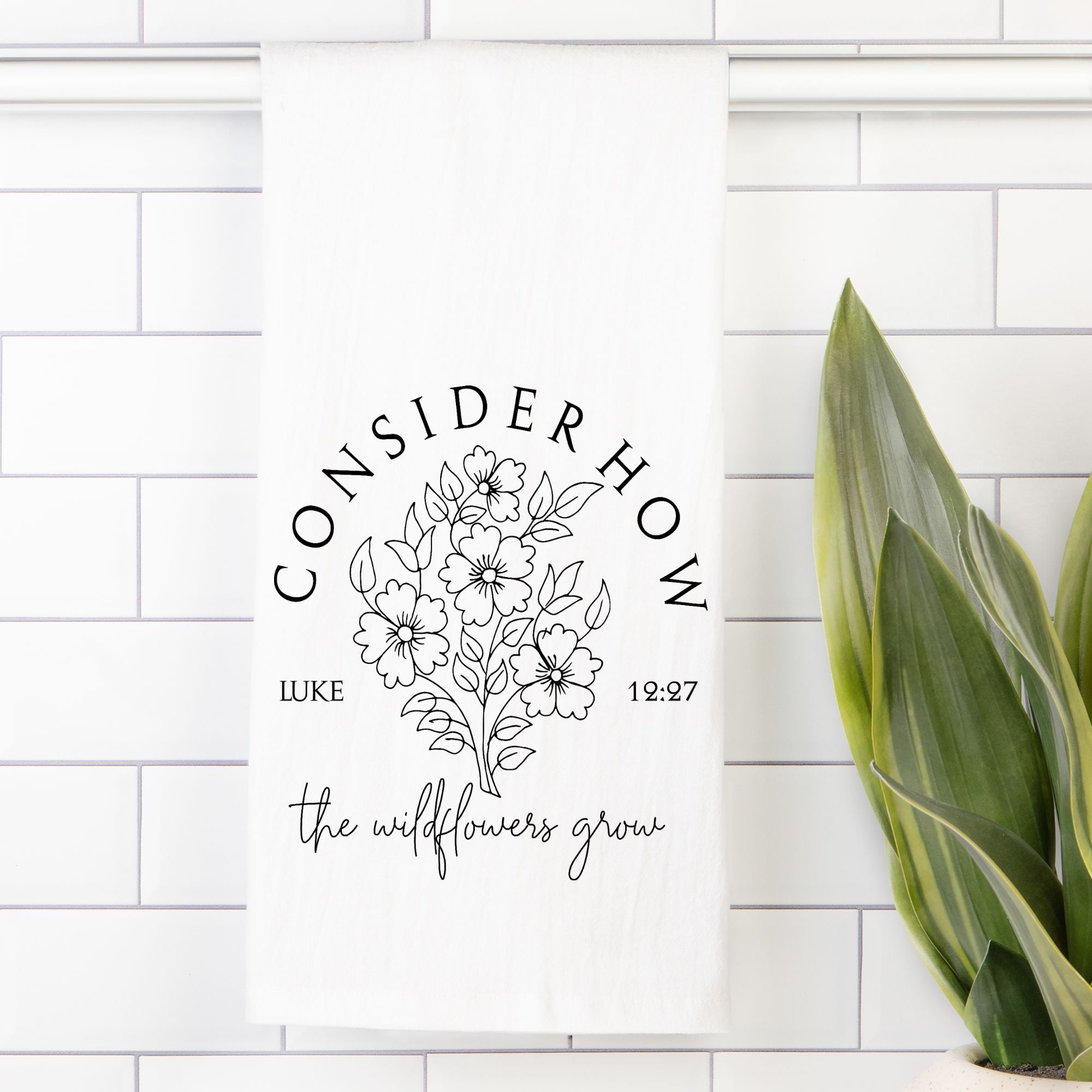 Decorative tea towel with floral design and text on a white tiled wall.