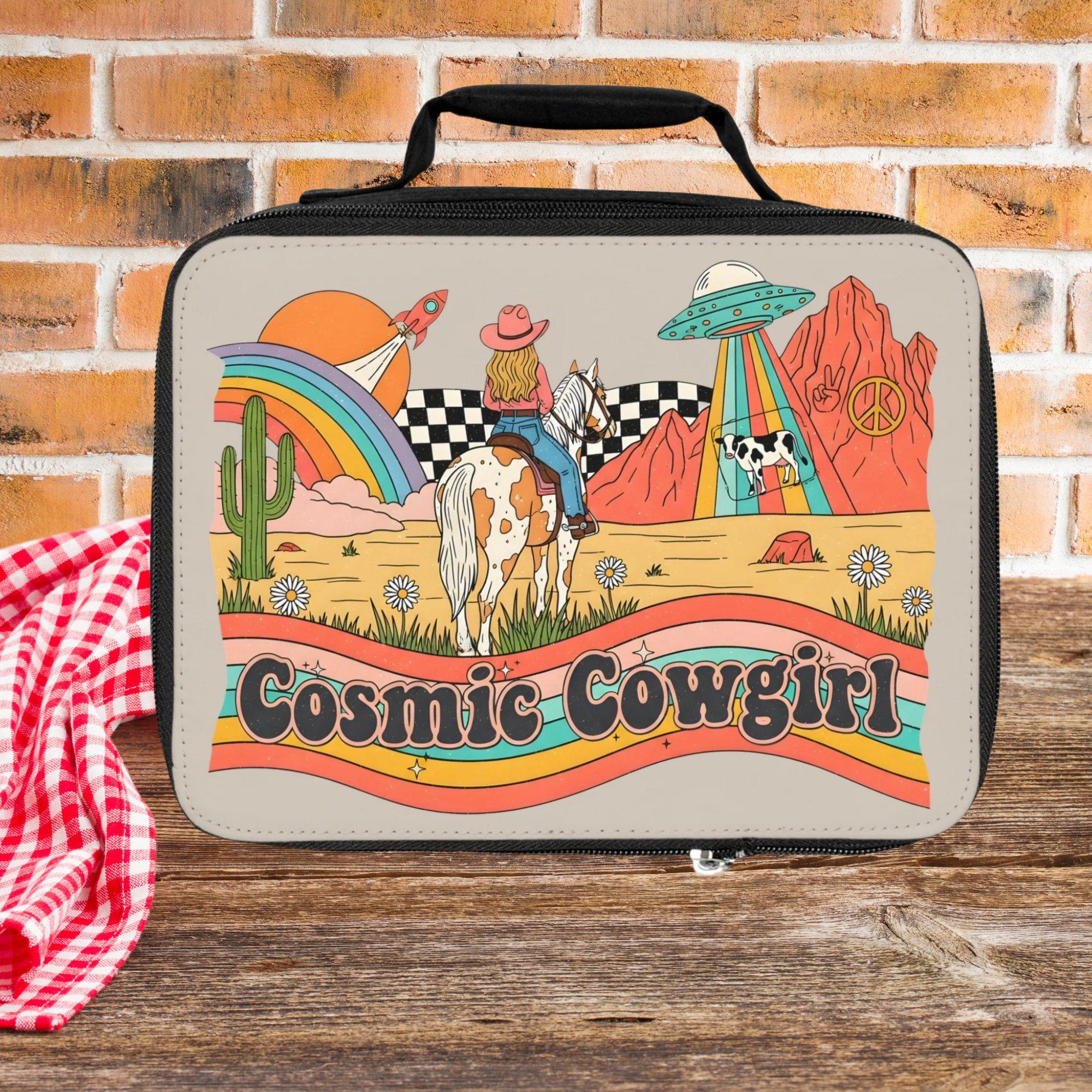 Lunch bag with 'Cosmic Cowgirl' design featuring a cowgirl, horse, and UFOs on a wooden picnic table and brick background.