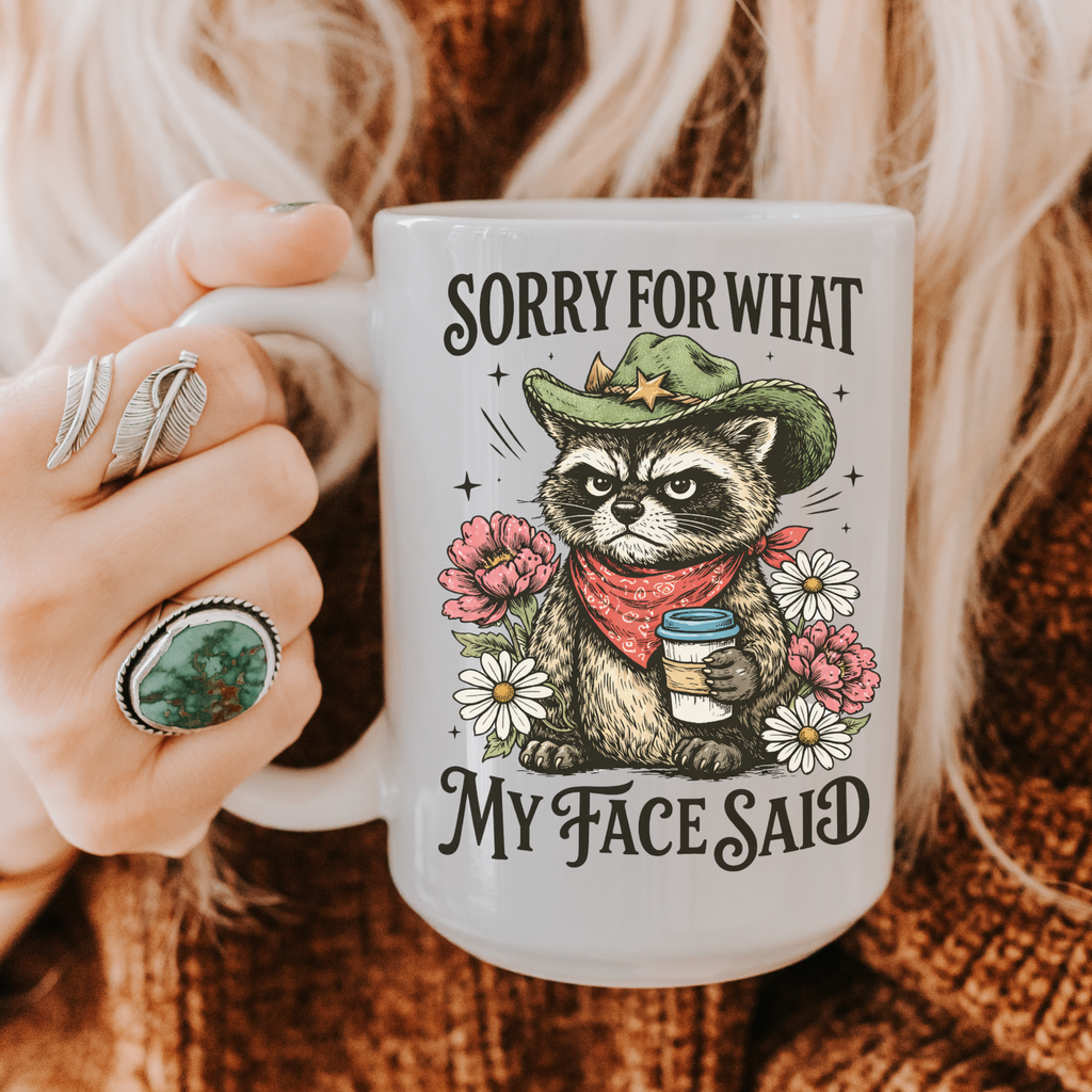 Mug with a cartoon raccoon holding a coffee cup, surrounded by flowers, and text 'Sorry for what my face said' held by a person.