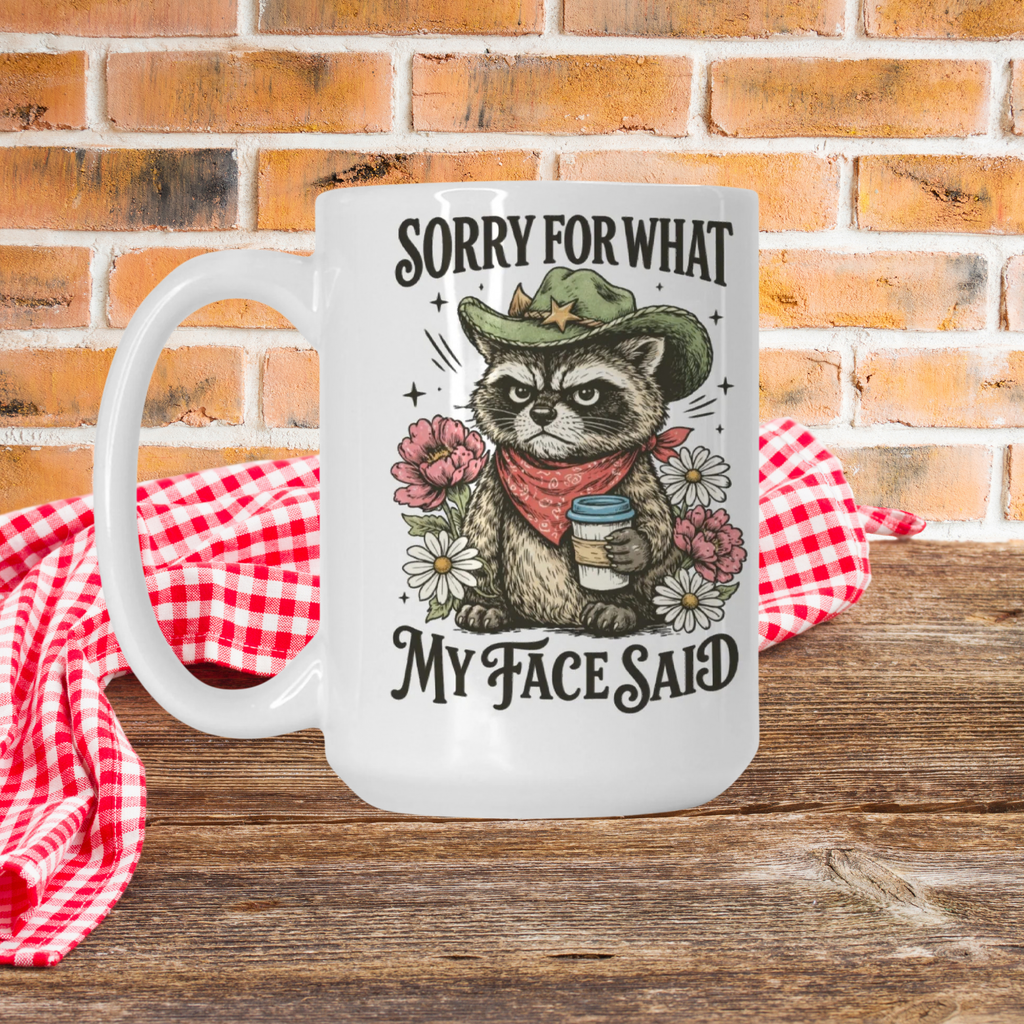White mug with a graphic of a raccoon wearing a cowboy hat and bandana, holding flowers, with text 'Sorry for what my face said' on a wooden picnic table with a red checkered cloth napkin and brick wall background.