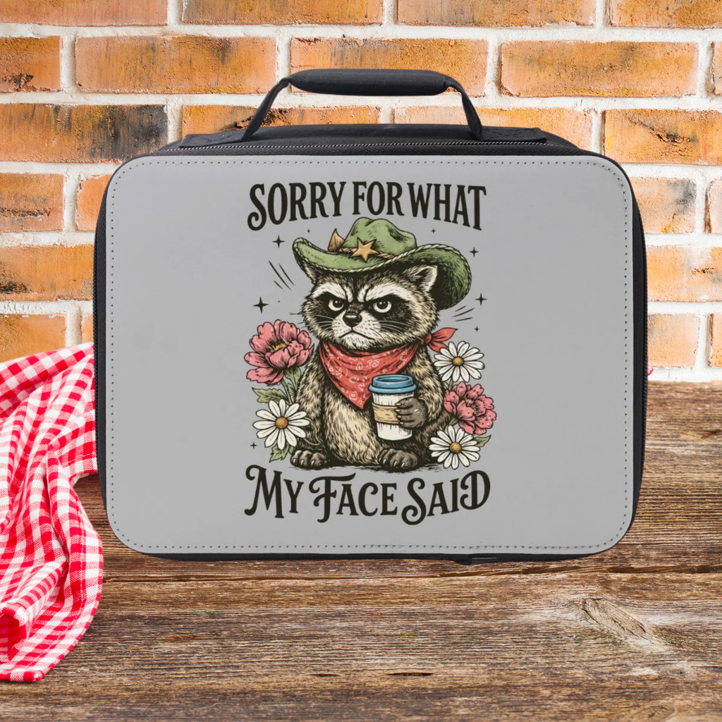 Black lunch bag with a cartoon raccoon holding a coffee cup and flowers, sitting on a wooden picnic table against a brick wall background.