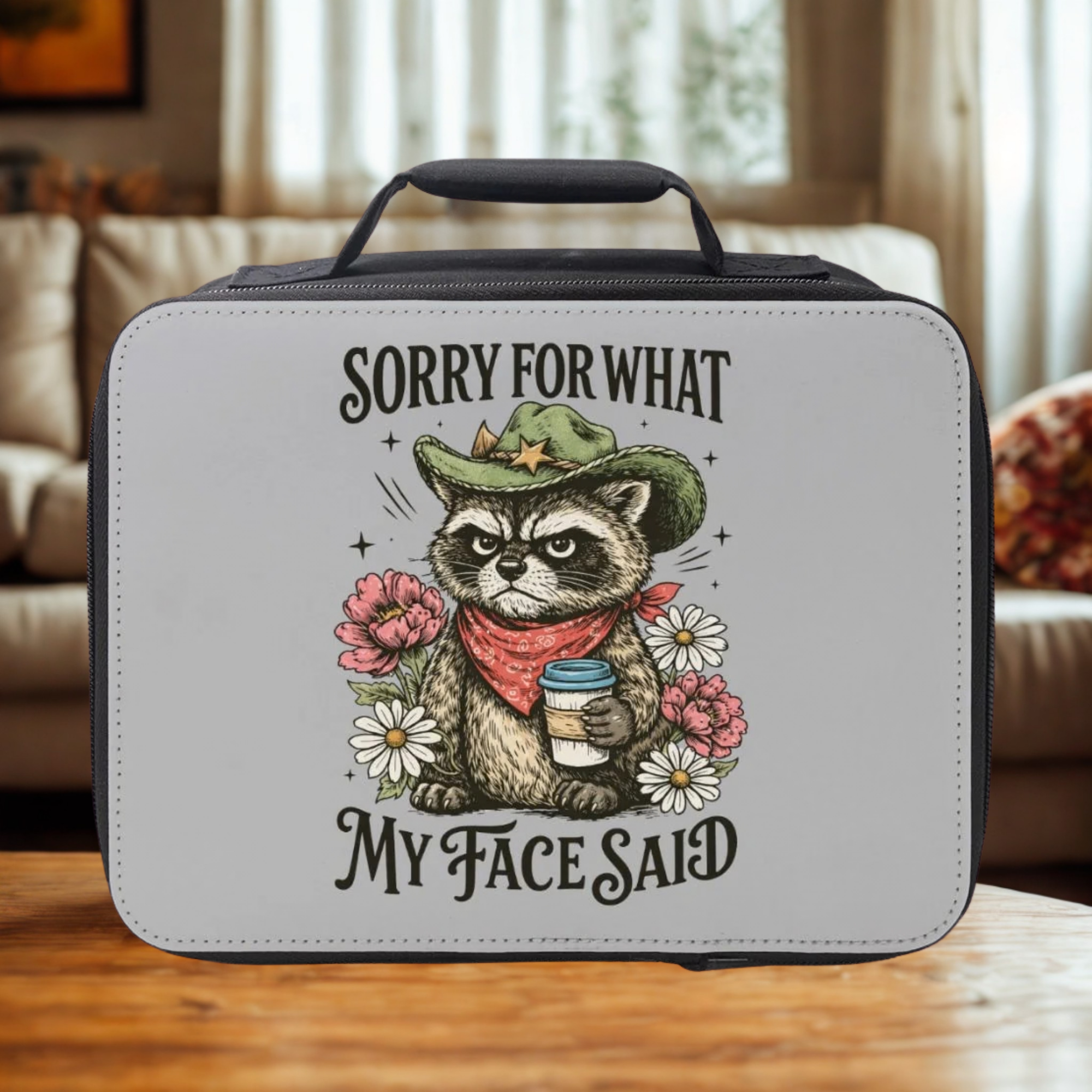 Gray lunch bag with a raccoon illustration and text 'Sorry For What My Face Said' on a wooden table in a cozy living room.