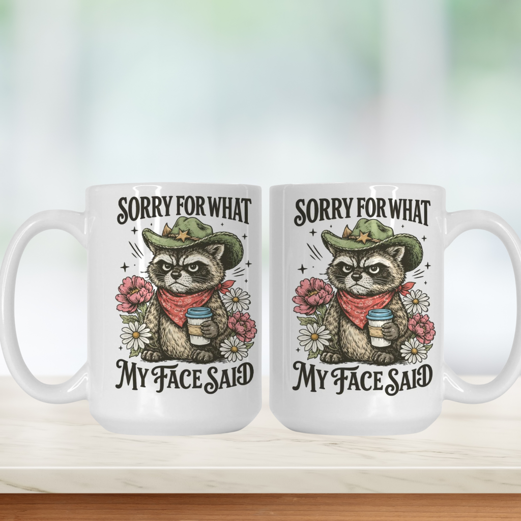 Two white 15oz. mugs with a cartoon raccoon wearing a cowboy hat and flowers, and text 'Sorry for what my face said' on a blurred background.