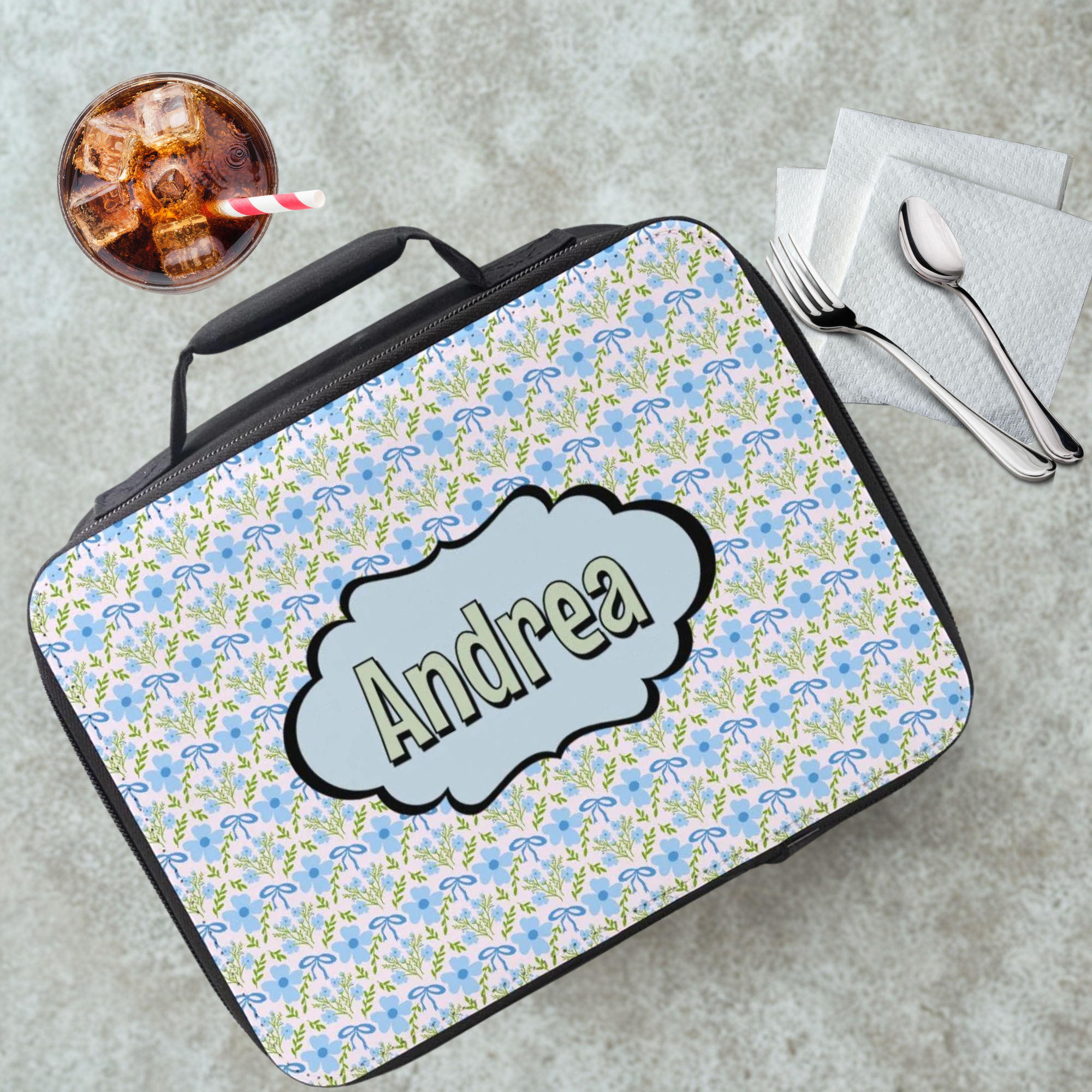 Floral-patterned lunch bag with 'Andrea' on a cafeteria table with a drink and utensils.