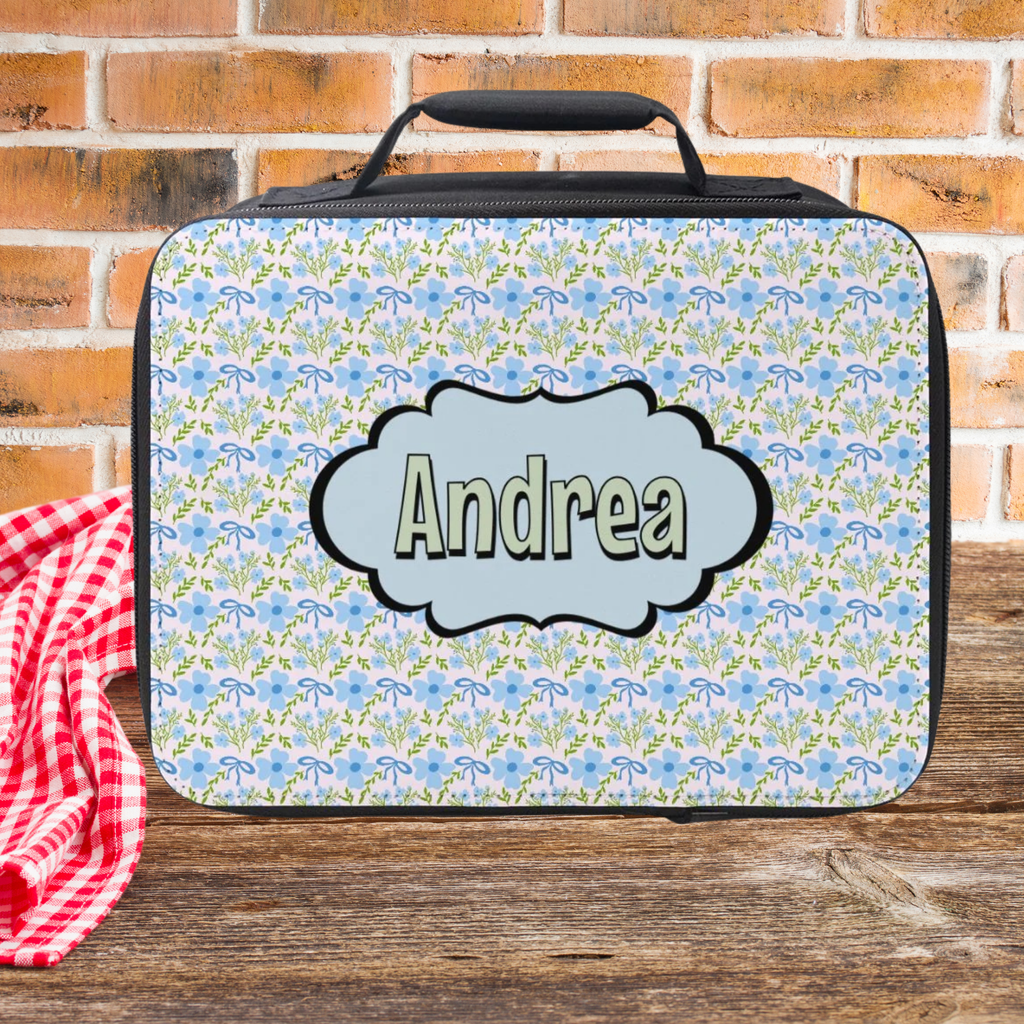 Floral patterned lunch bag with 'Andrea' on a wooden picnic table and brick wall background