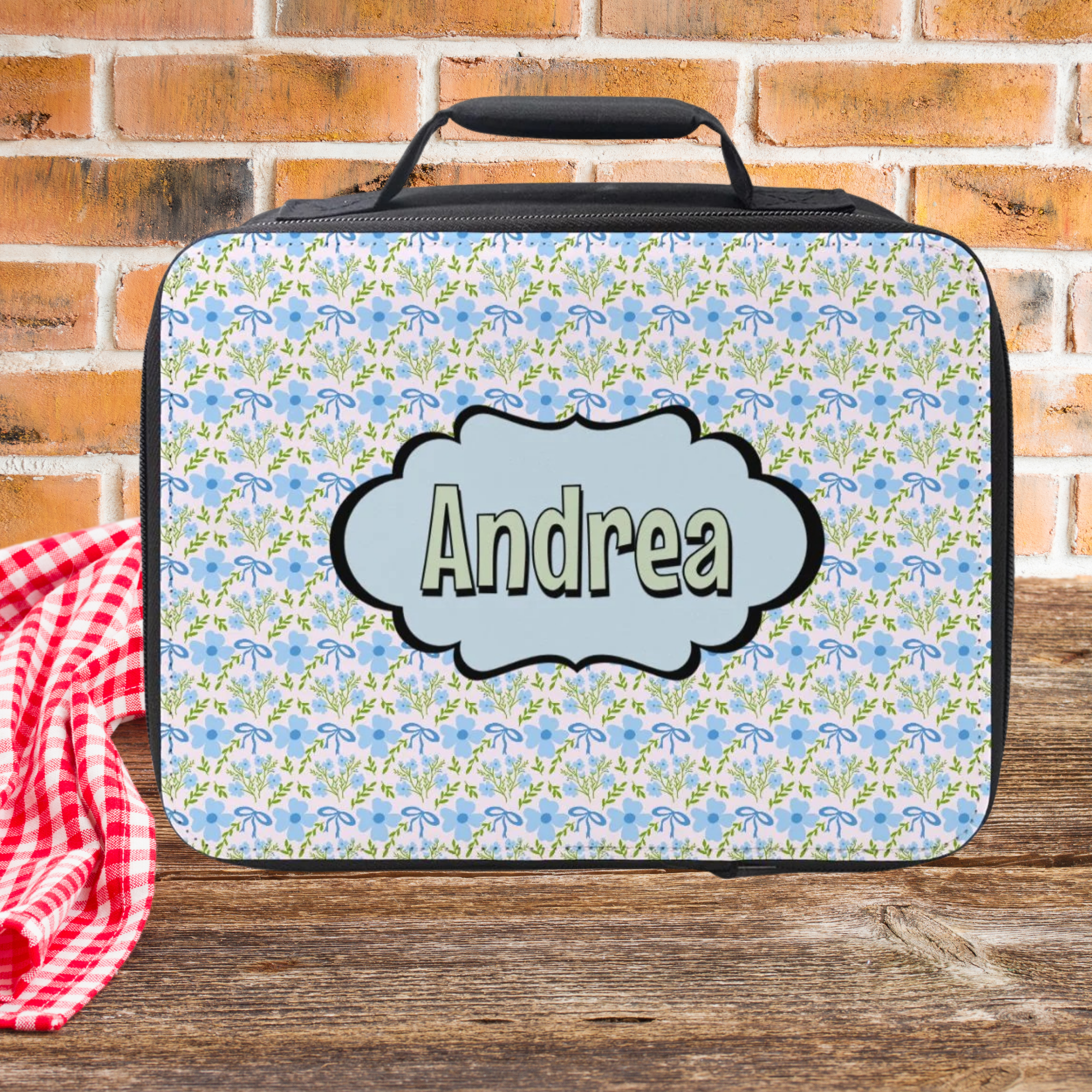 Floral patterned lunch bag with 'Andrea' on a wooden picnic table and brick wall background