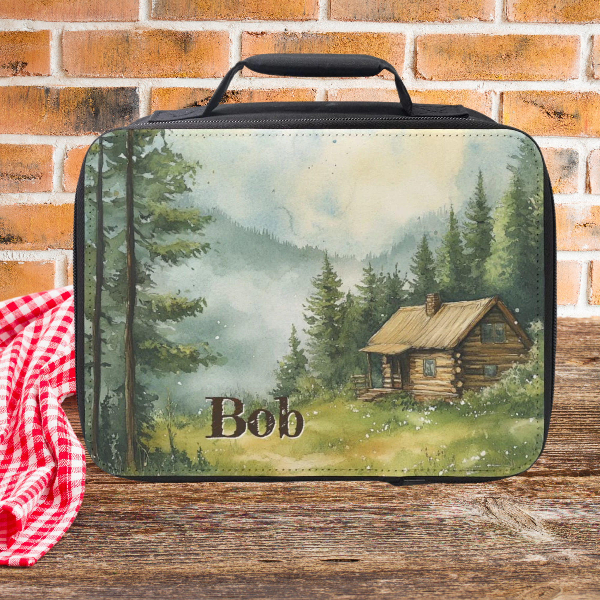  insulated lunch bag with a cabin design and 'Bob' text, placed on a wooden picnic table with a brick wall background.