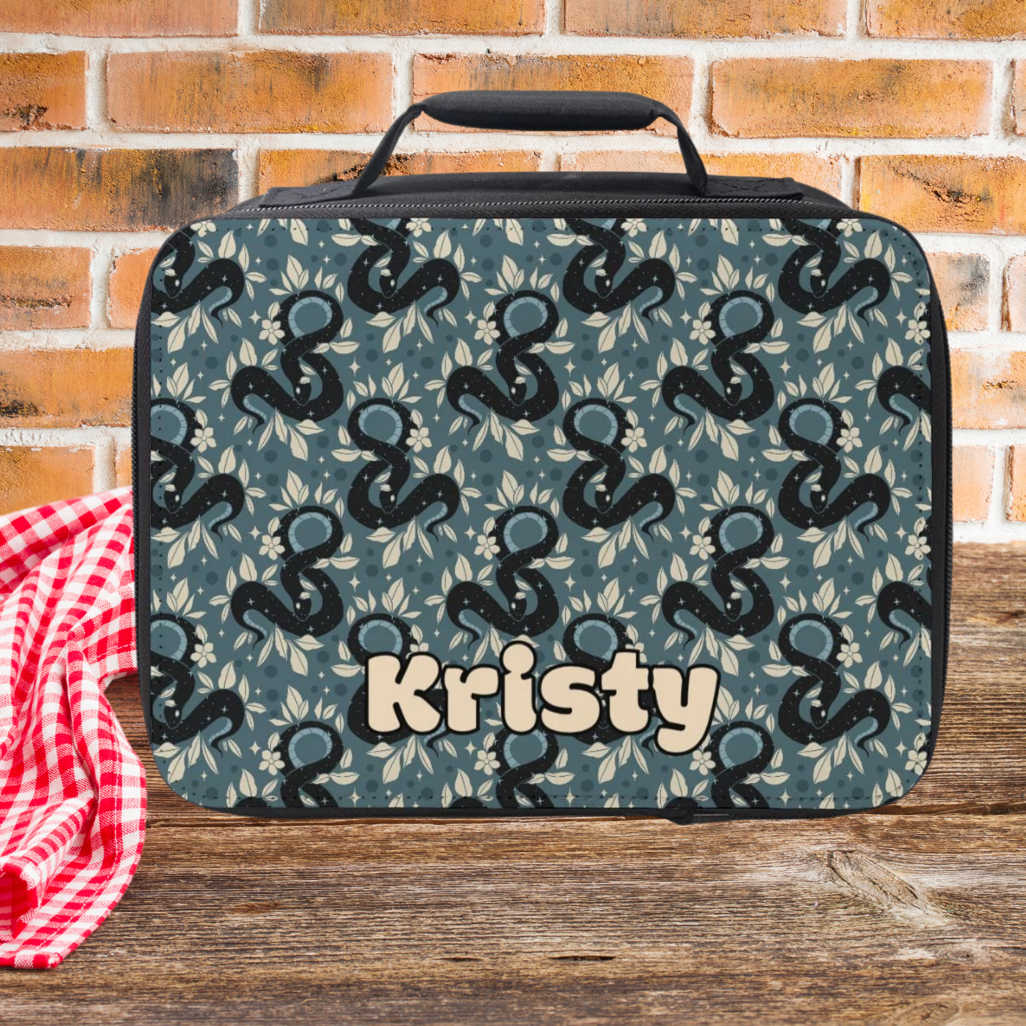 Lunch bag with snake pattern and 'Kristy' custom text on a wooden picnic table with brick background