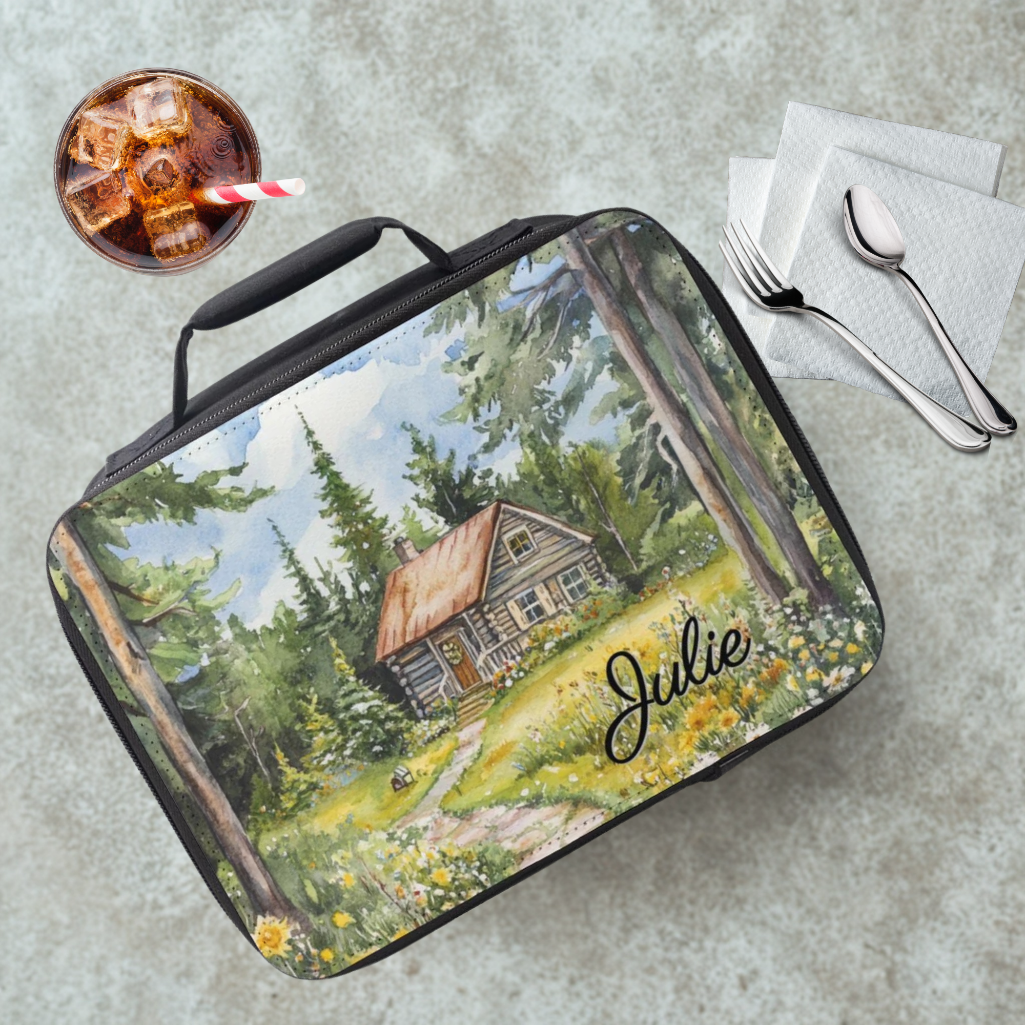 Lunch bag with a forest scene and cabin design, personalized with 'Julie', on a cafeteria table with a drink and utensils.