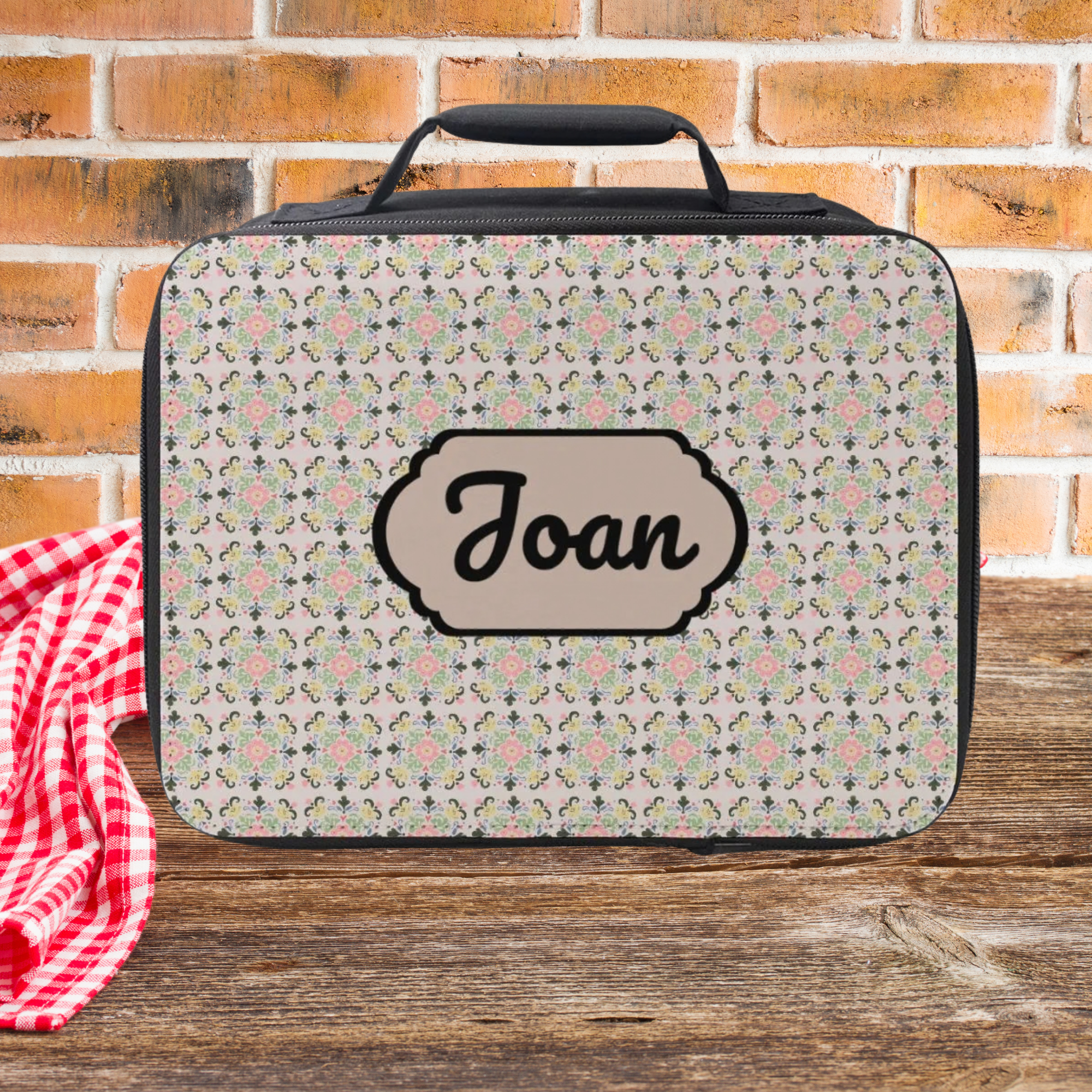 Floral patterned lunch bag with 'Joan' custom text on a wooden picnic table and brick wall background