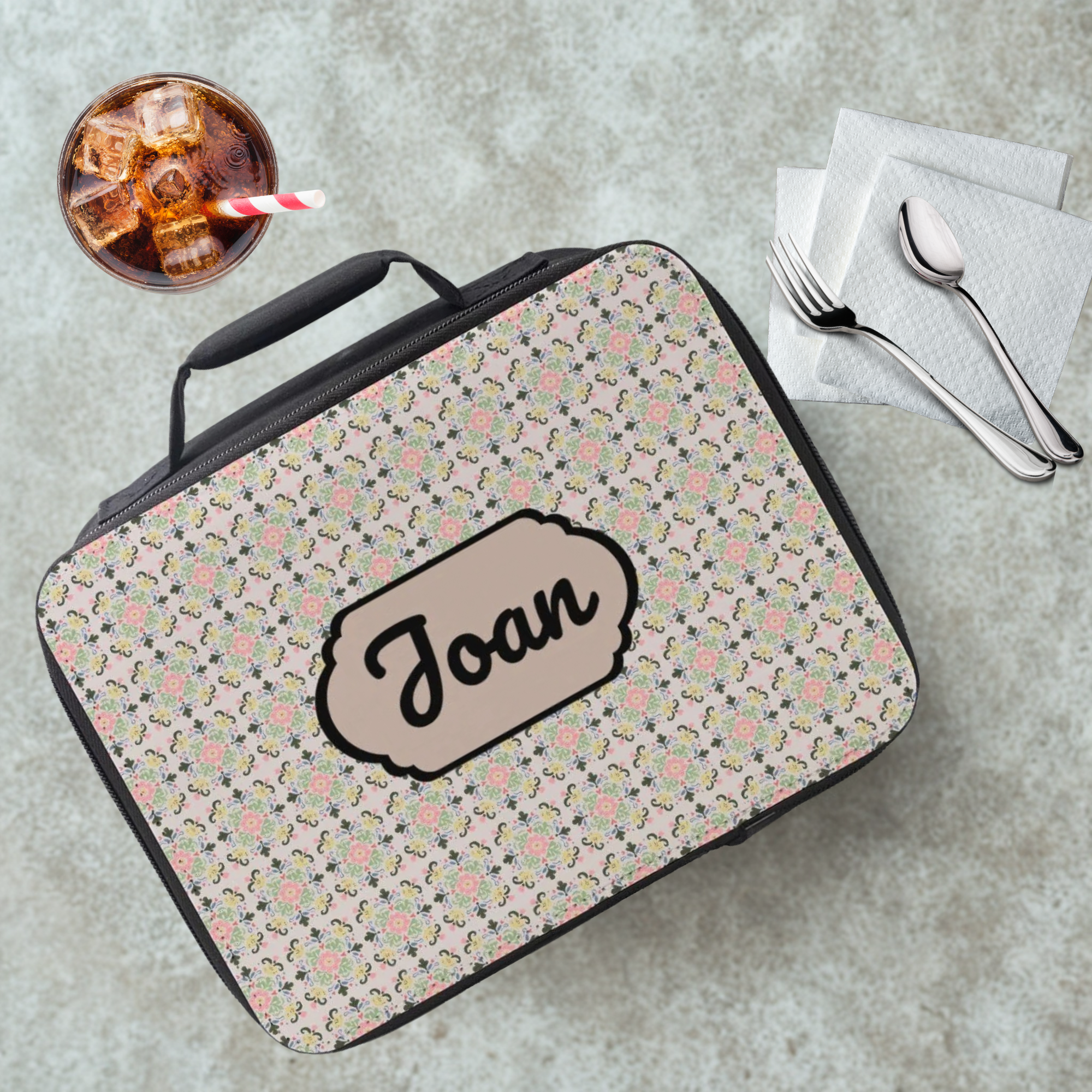 Floral patterned lunch bag with 'Joan' custom text on a cafeteria table with a drink and utensils.