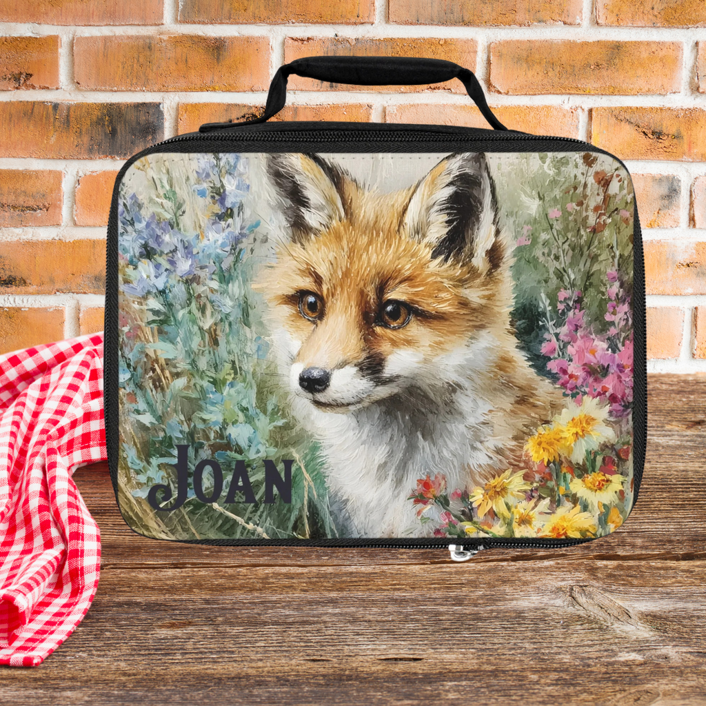 Lunch bag with a fox design and 'JOAN' personalized text on a wooden picnic table with a brick wall background.