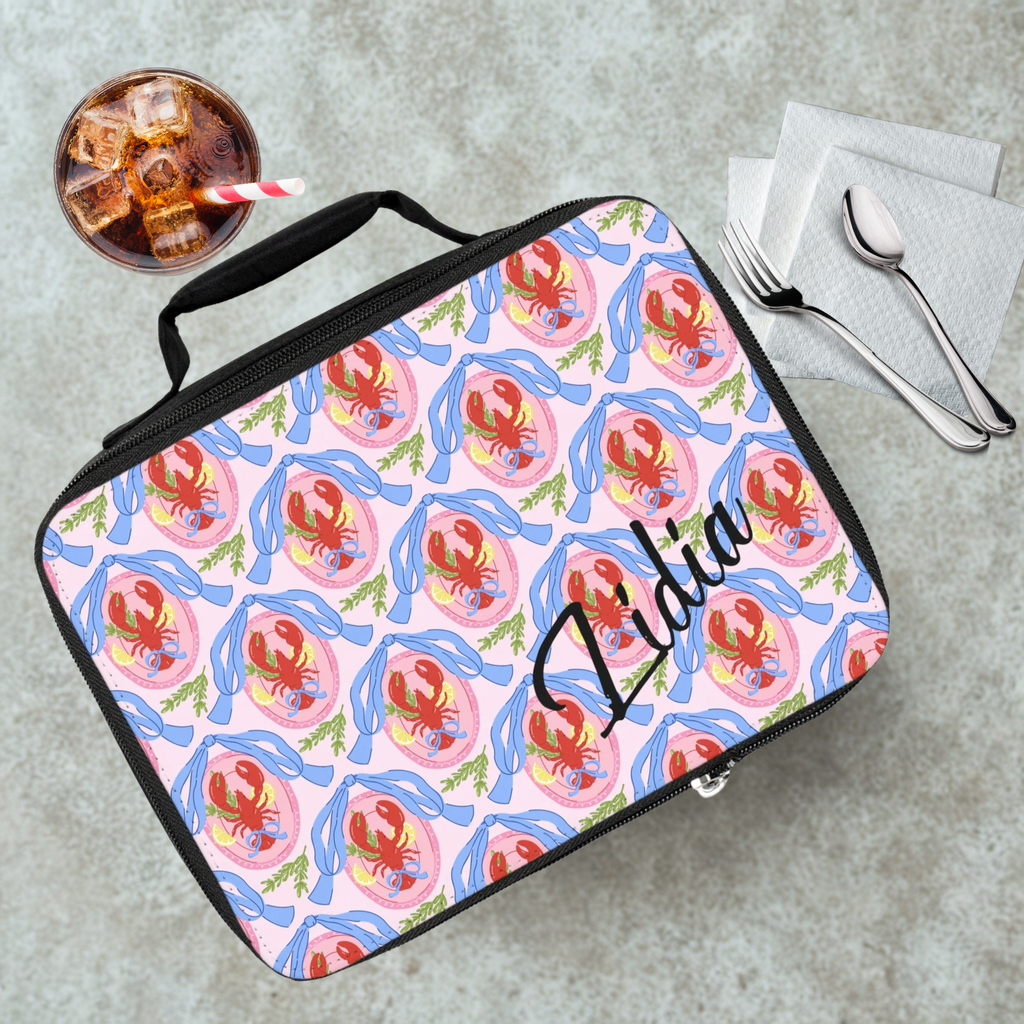 Lunch bag with lobster pattern and 'Lidia' personalized text on a cafeteria table with a drink and utensils.