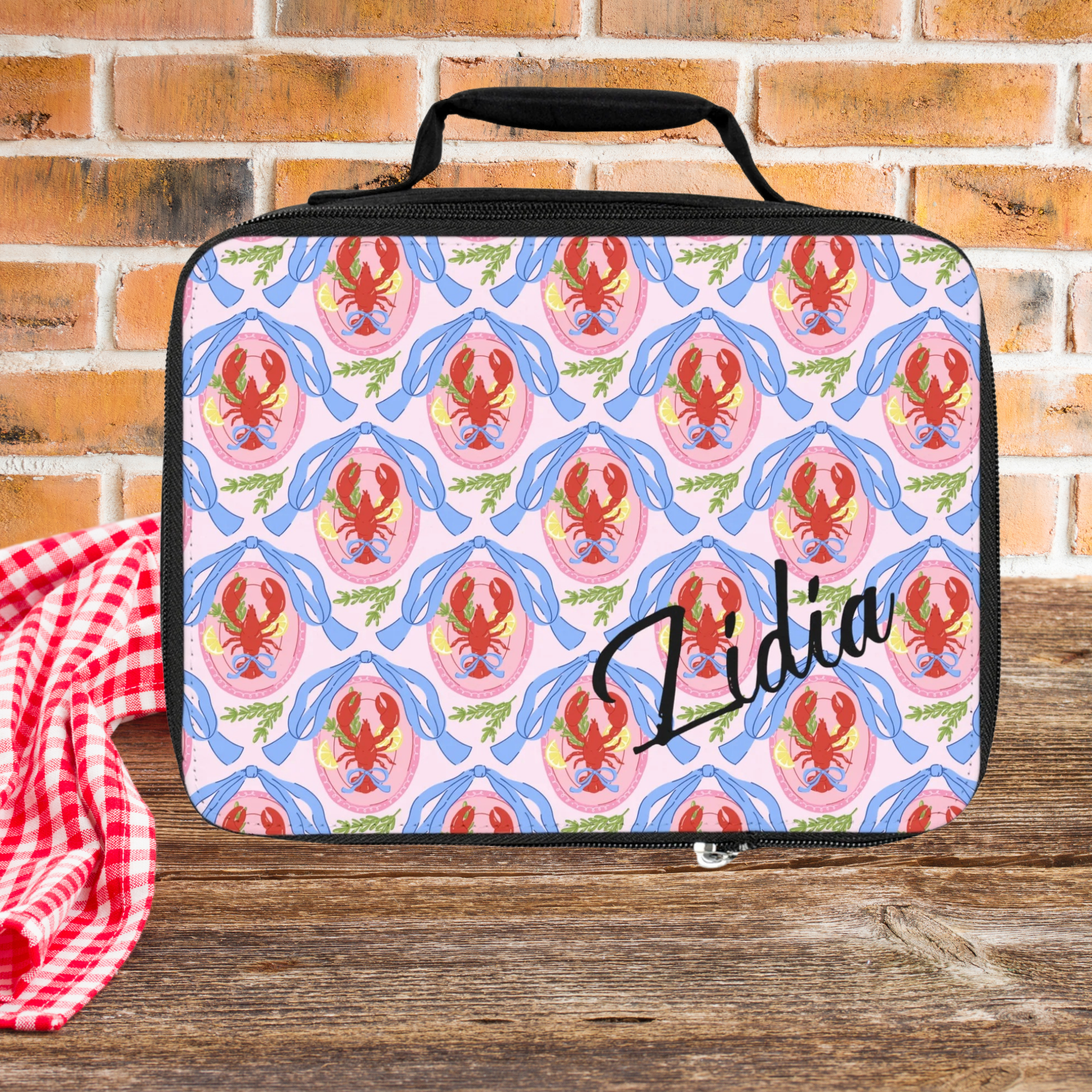  insulated lunch bag with lobster pattern and 'Lidia' custom text on a wooden picnic table with brick background