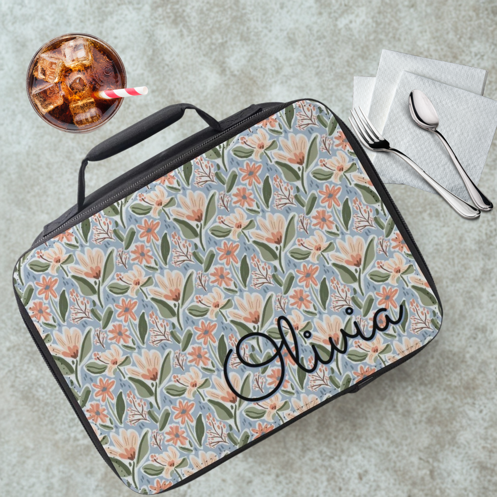 Floral patterned lunch bag with 'Olivia' on a cafeteria table with a drink and utensils.