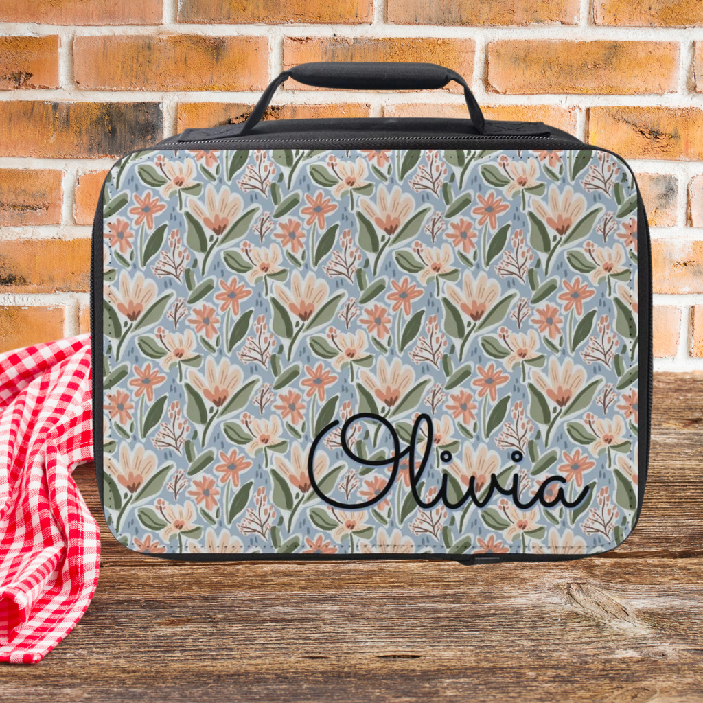 Floral-patterned lunch bag with 'Olivia' custom text on a wooden picnic table and brick wall background