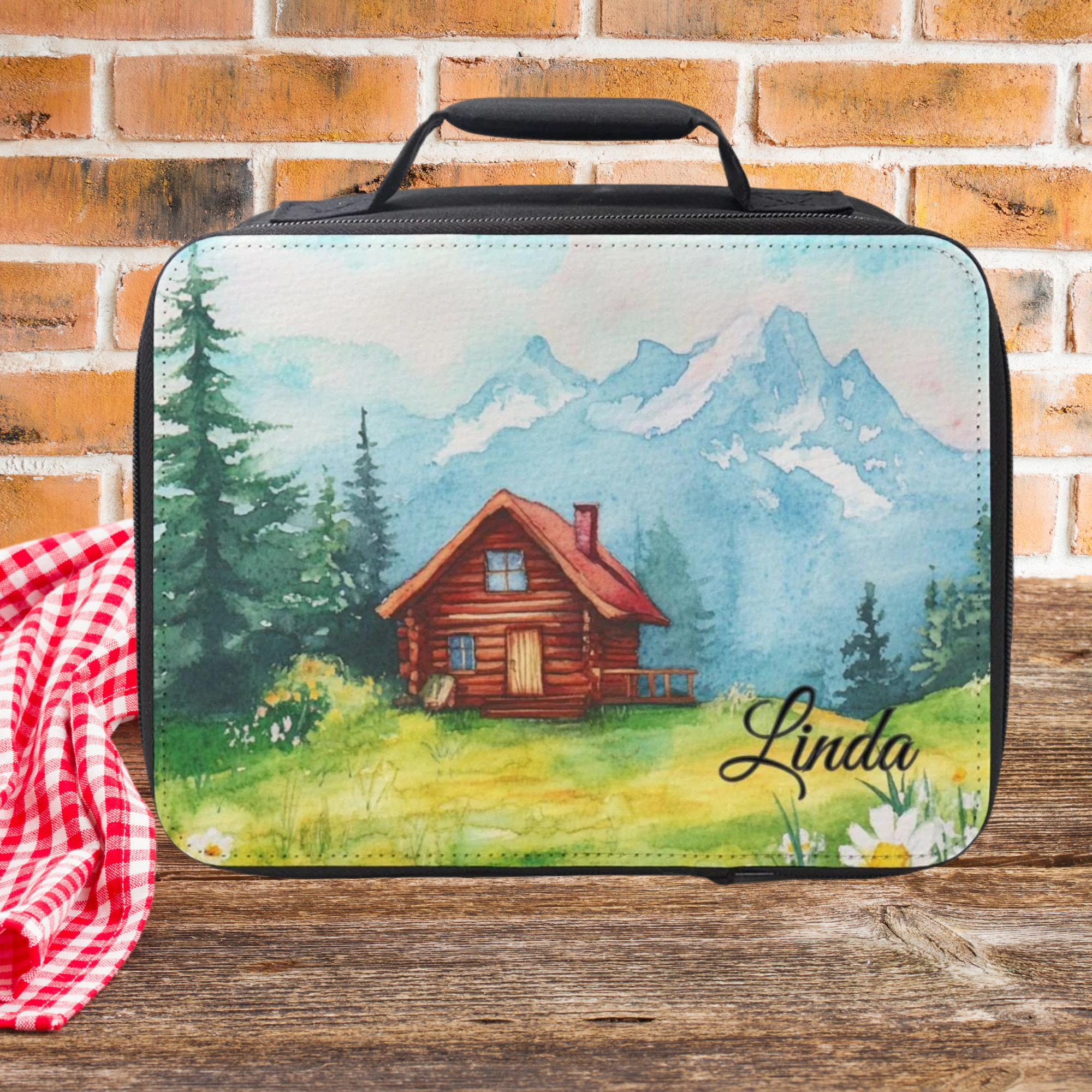 Lunch bag with a scenic design of a cabin and mountains, placed on a wooden surface with a brick wall background.