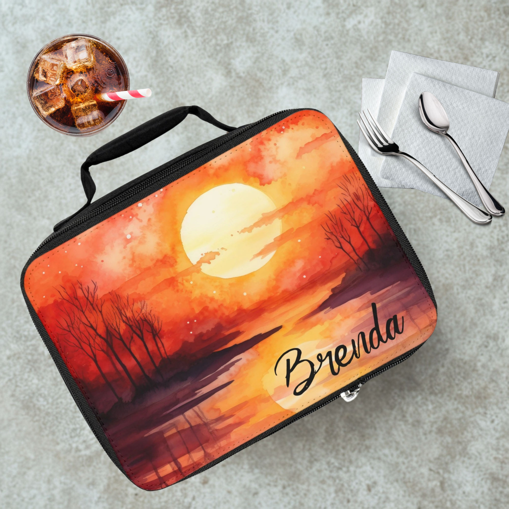 Lunch bag with sunset design and 'Brenda' text on a cafeteria table with a drink and utensils.