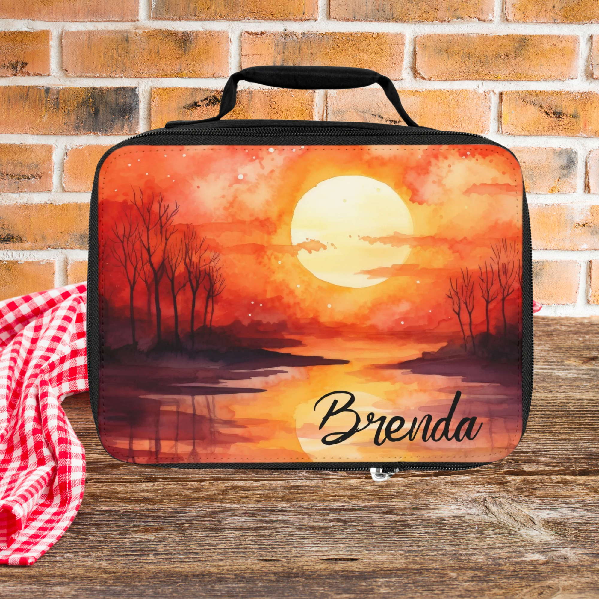  insulated lunch bag with sunset design and 'Brenda' text on a wooden picnic table with brick wall background.