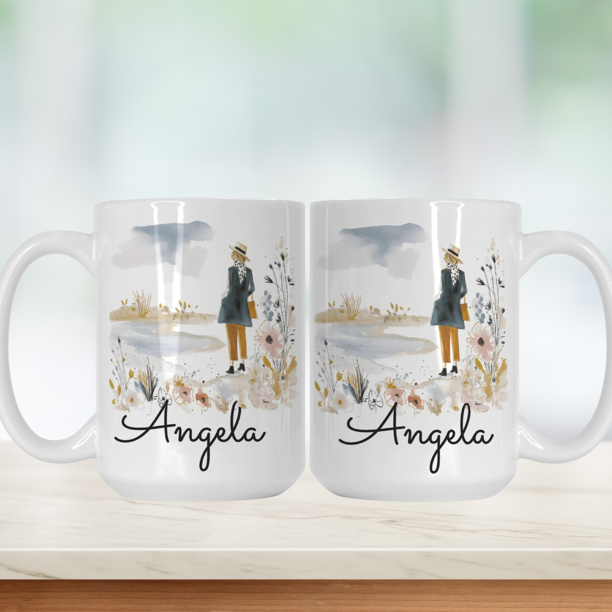 Two white mugs with a design of a person holding an umbrella, flowers, and the name 'Angela' on a light background.