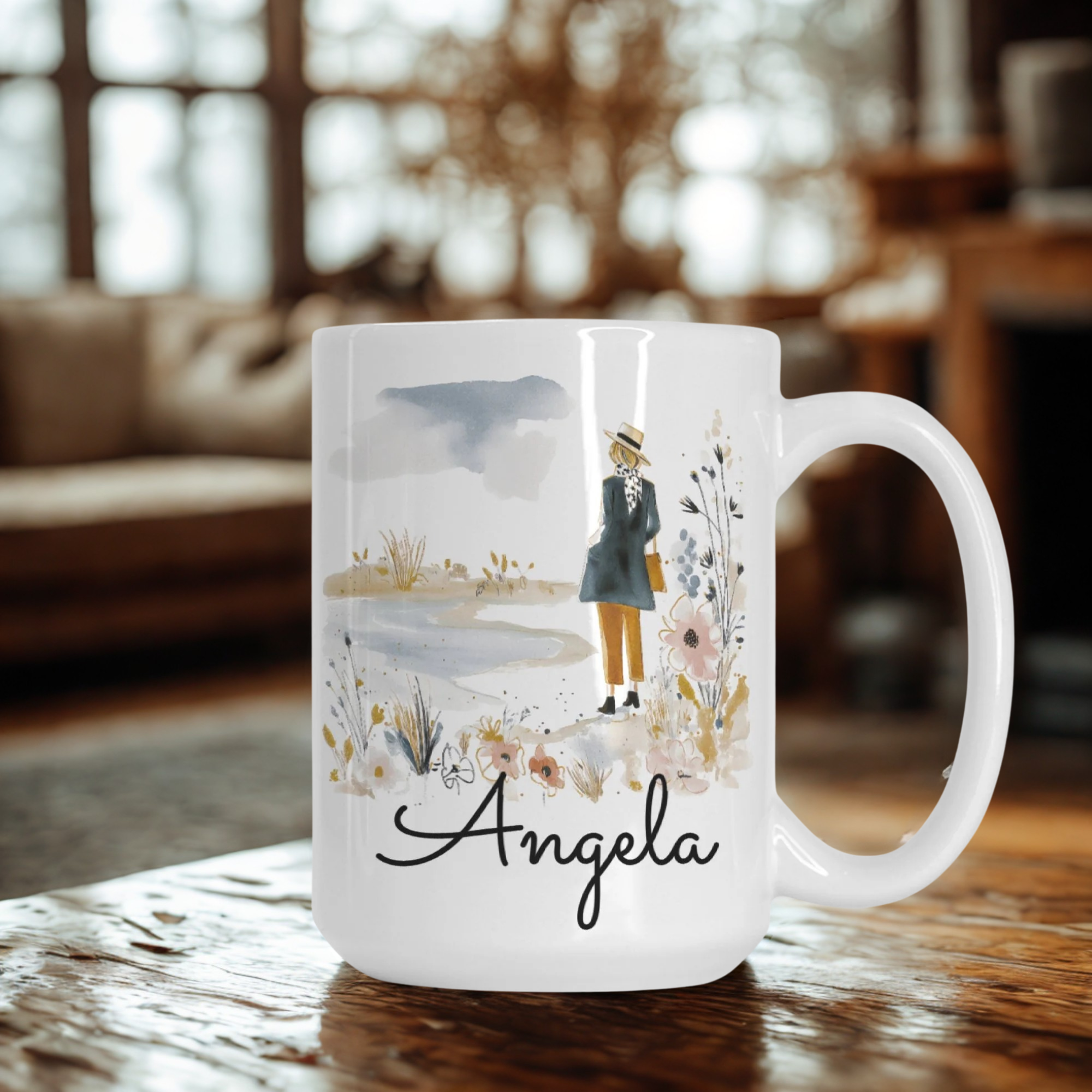 White mug with a design and the name 'Angela' on a wooden table in a cozy room.