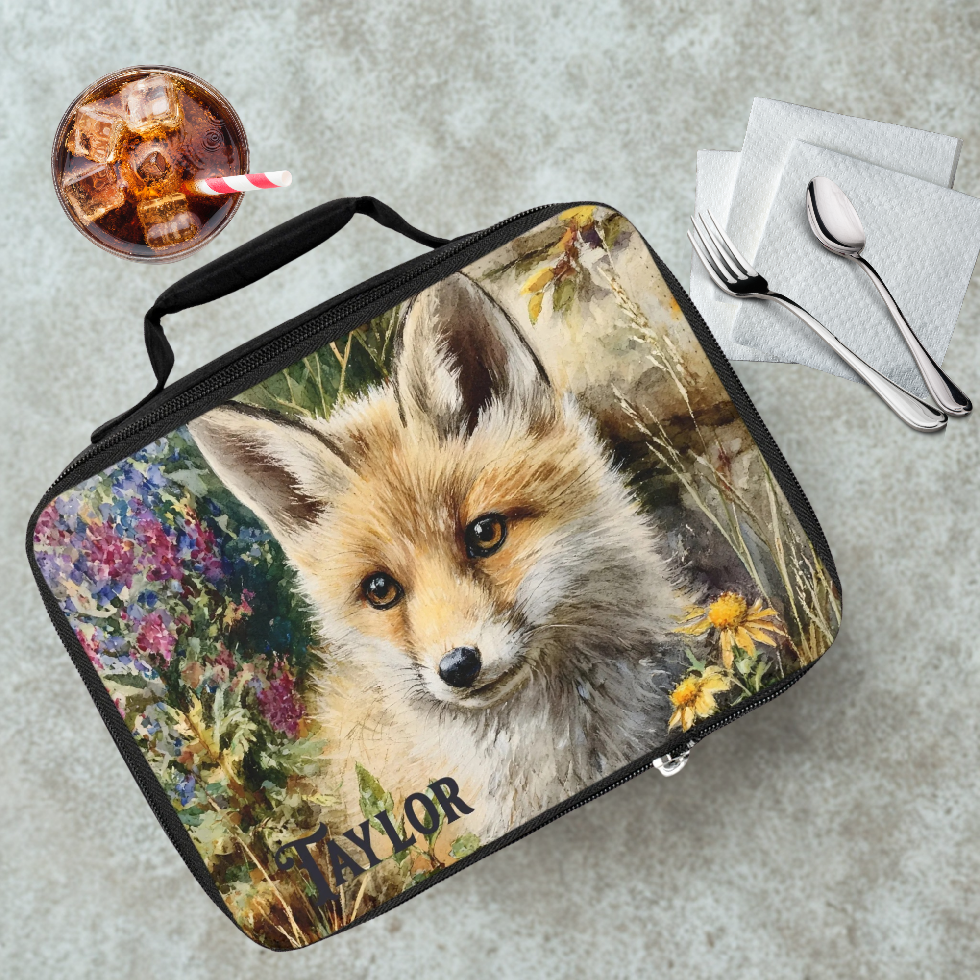 Lunch bag with a fox design and the name 'Taylor' on a cafeteria table with a drink and utensils.