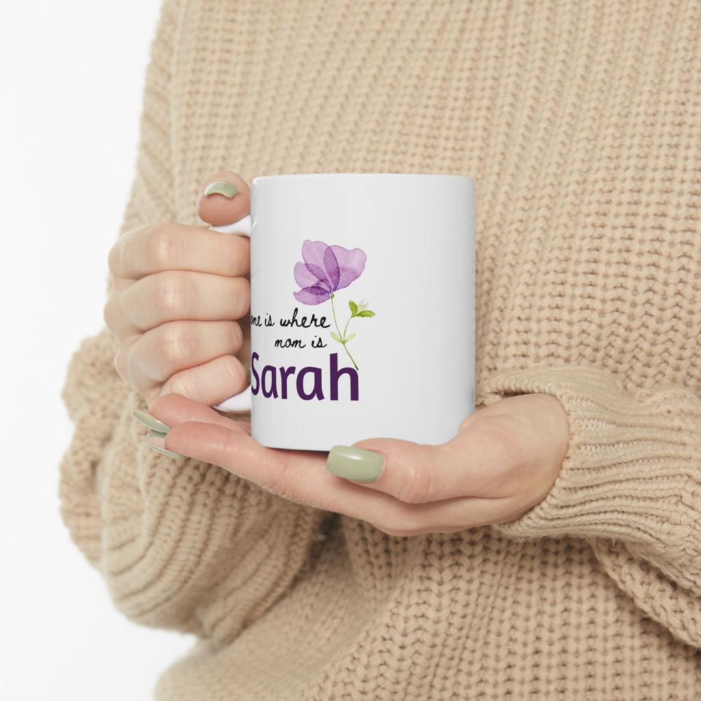 Person holding an 11oz mug with a floral design and text 'Home Is Where Mom Is', wearing a beige sweater.
