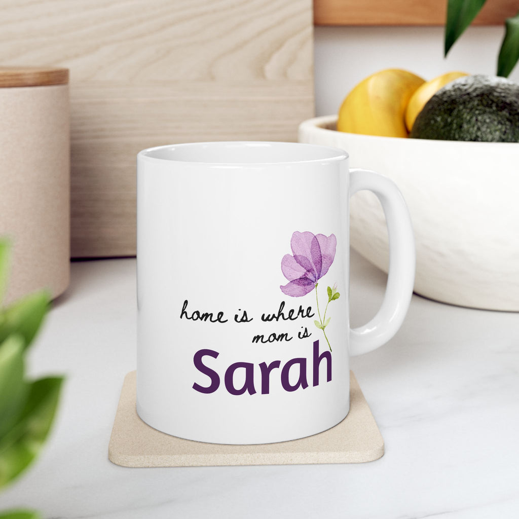 White 11oz mug with floral design and text 'Home Is Where Mom Is' on a coaster, placed on a surface with a plant and container in the background.
