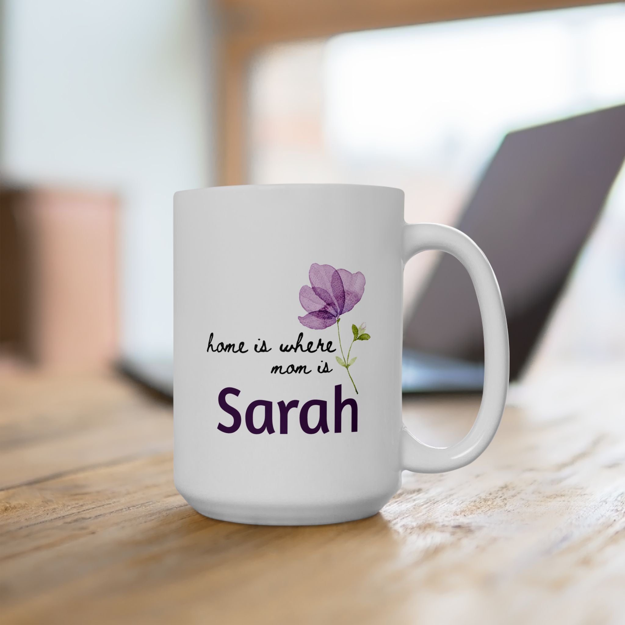 White 15oz mug with purple flower design and text 'Home Is Where Mom Is' on a wooden desk with blurred office in the background.