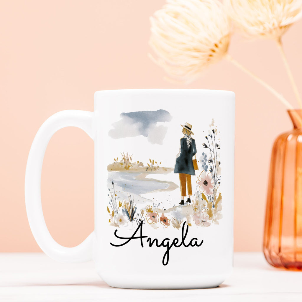 White mug with a design of a person standing by a lake, flowers, and the name 'Angela' on a peach background.