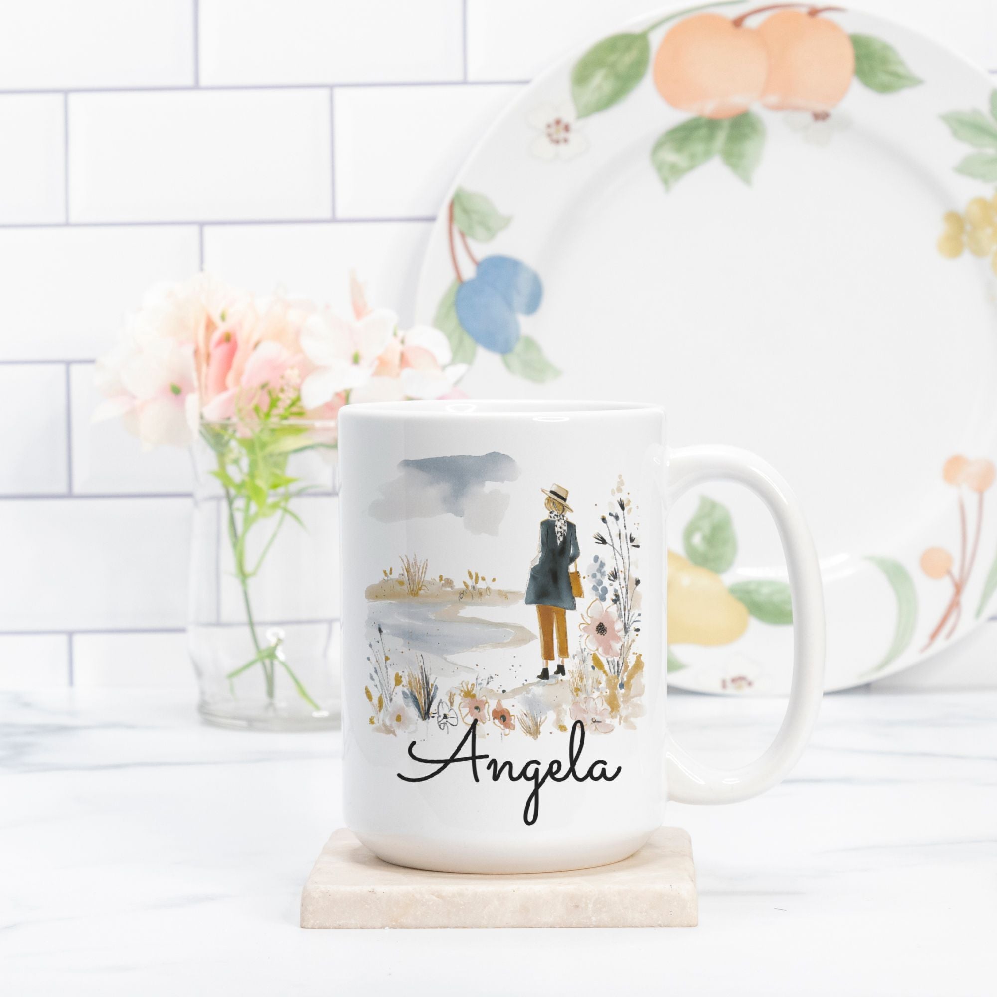 Mug with a scenic design and 'Angela' text, placed on a white surface with a decorative plate in the background.