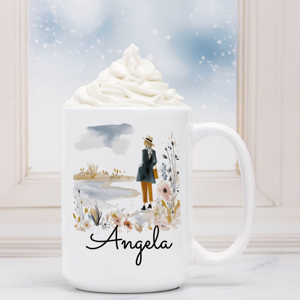 White mug with a design of a person in a coat and hat, flowers, and the name 'Angela'. Behind the mug is a steaming cup of hot chocolate with whipped cream.