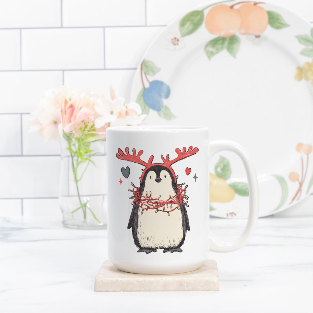 Mug with a penguin design wearing reindeer antlers on a white surface.