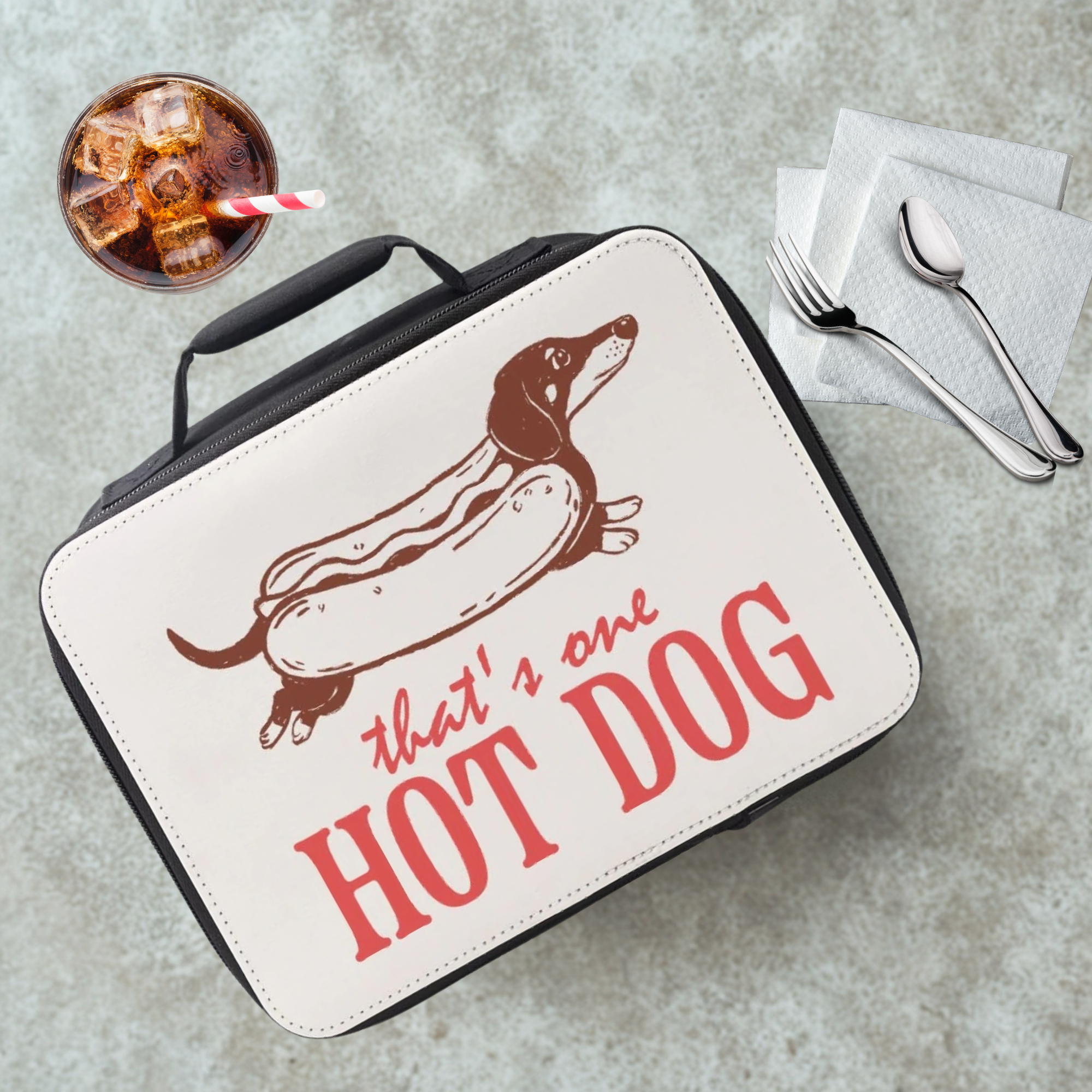 Lunch bag with a dachshund as a hot dog graphic and text 'that's one HOT DOG' on a cafeteria table with a drink and utensils.