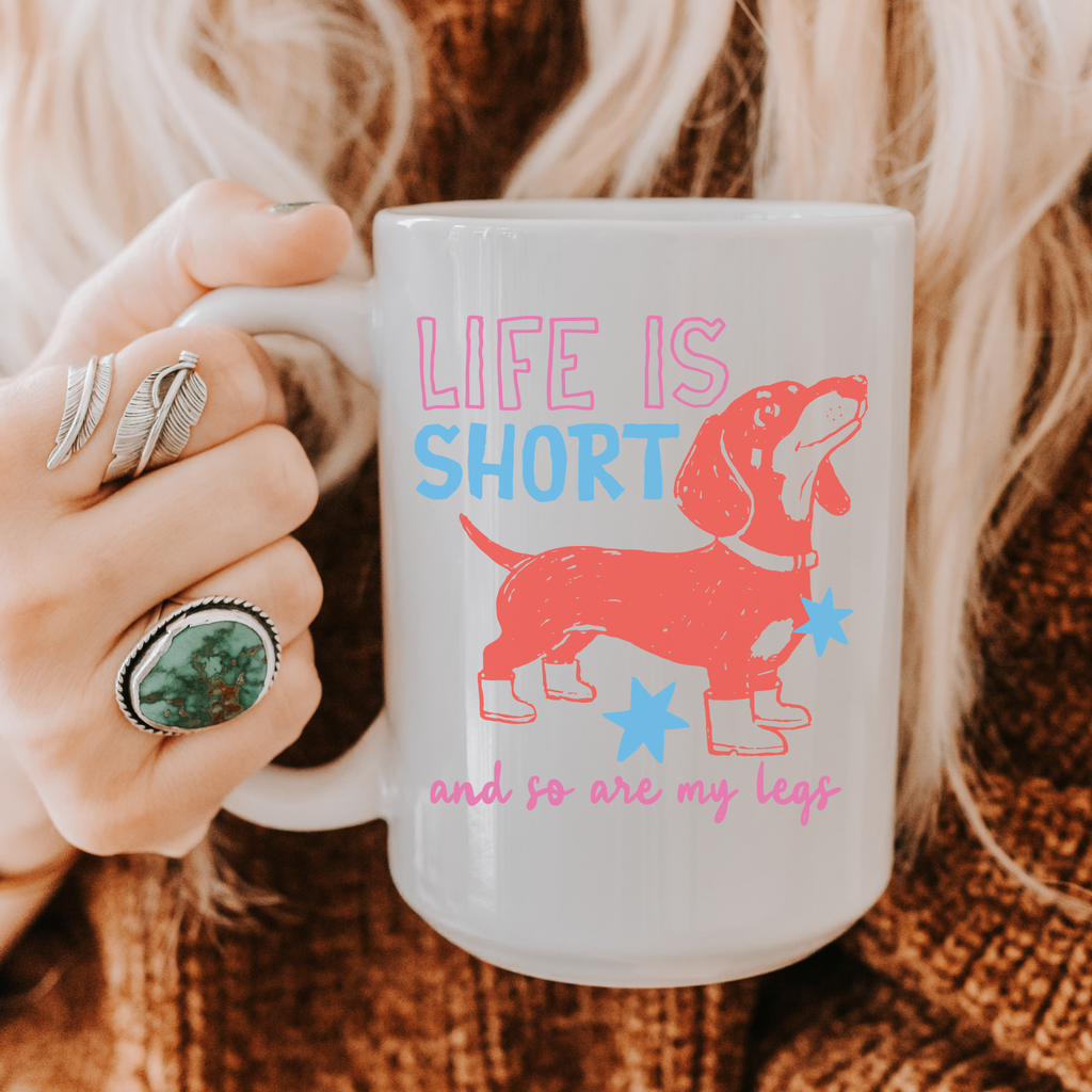Person holding a 15oz. mug with a dachshund design and text 'Life is short and so are my legs' on a textured surface.