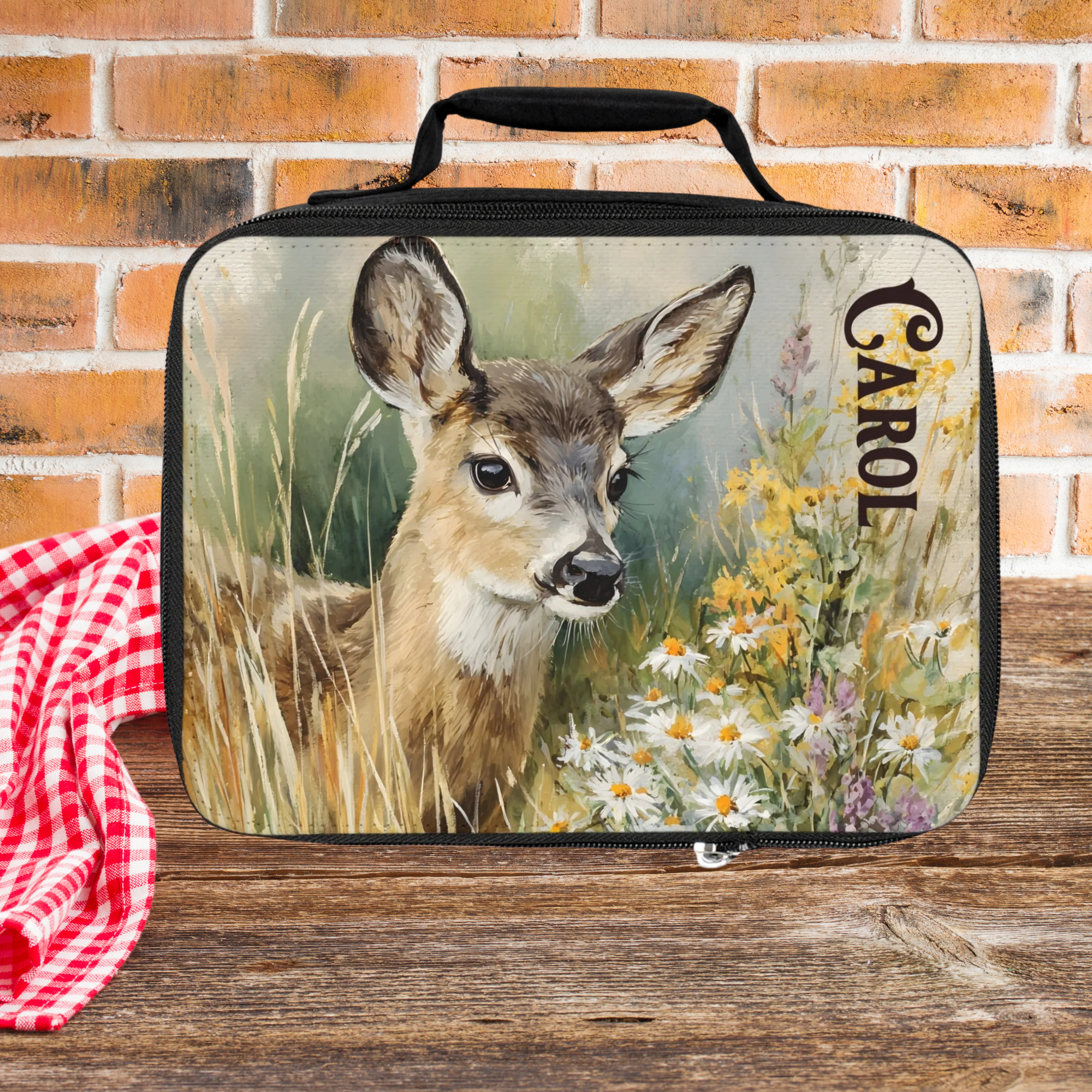 Lunch bag with a deer design and 'Carol' text on a wooden picnic table with a brick wall background.
