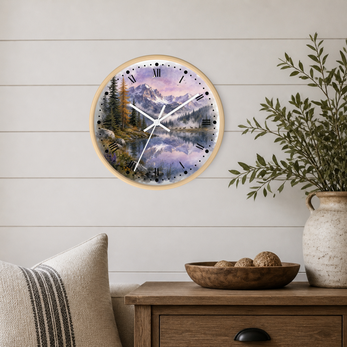 A round wall clock with a light wood frame displays a scenic mountain landscape with a lake and pine trees. The clock is mounted on a white shiplap wall above a wooden side table with a bowl, decorative vase, and cushion.