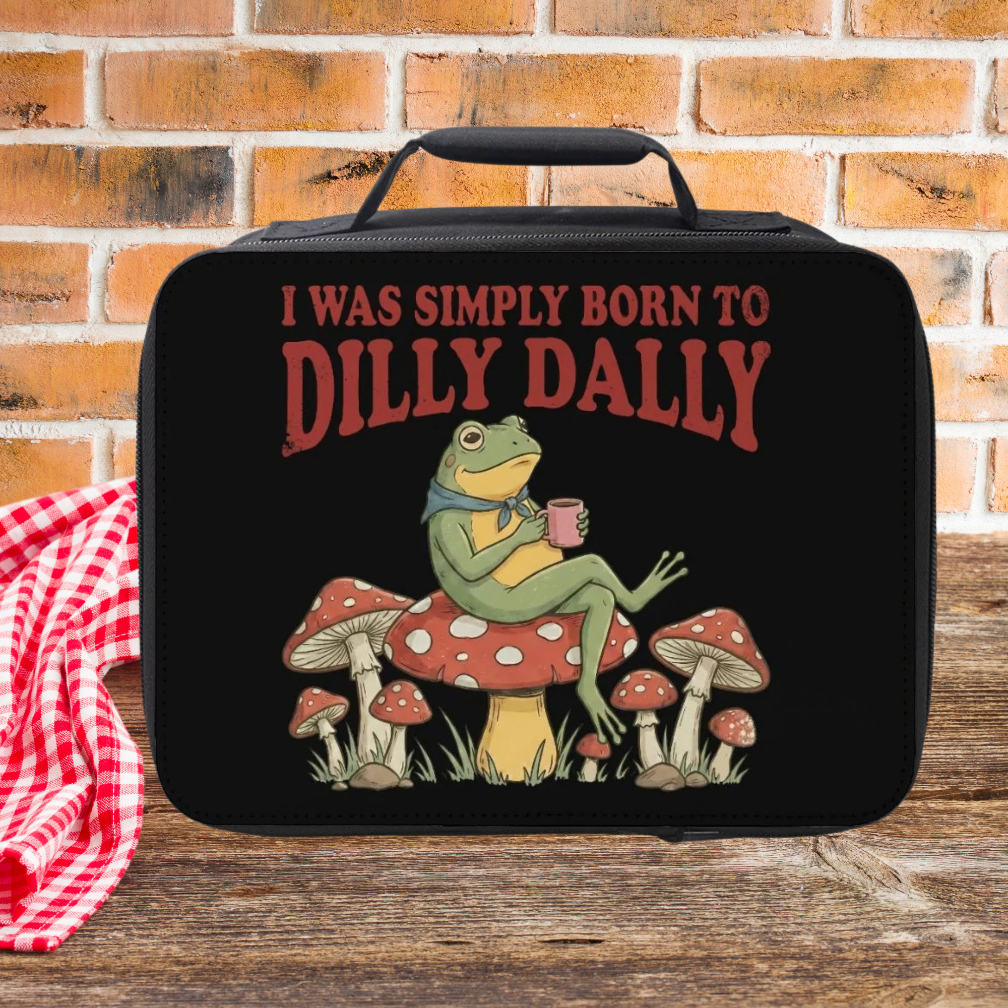 Black lunch bag with a frog graphic and text 'I was simply born to dilly dally' on a wooden picnic table against a brick wall background.