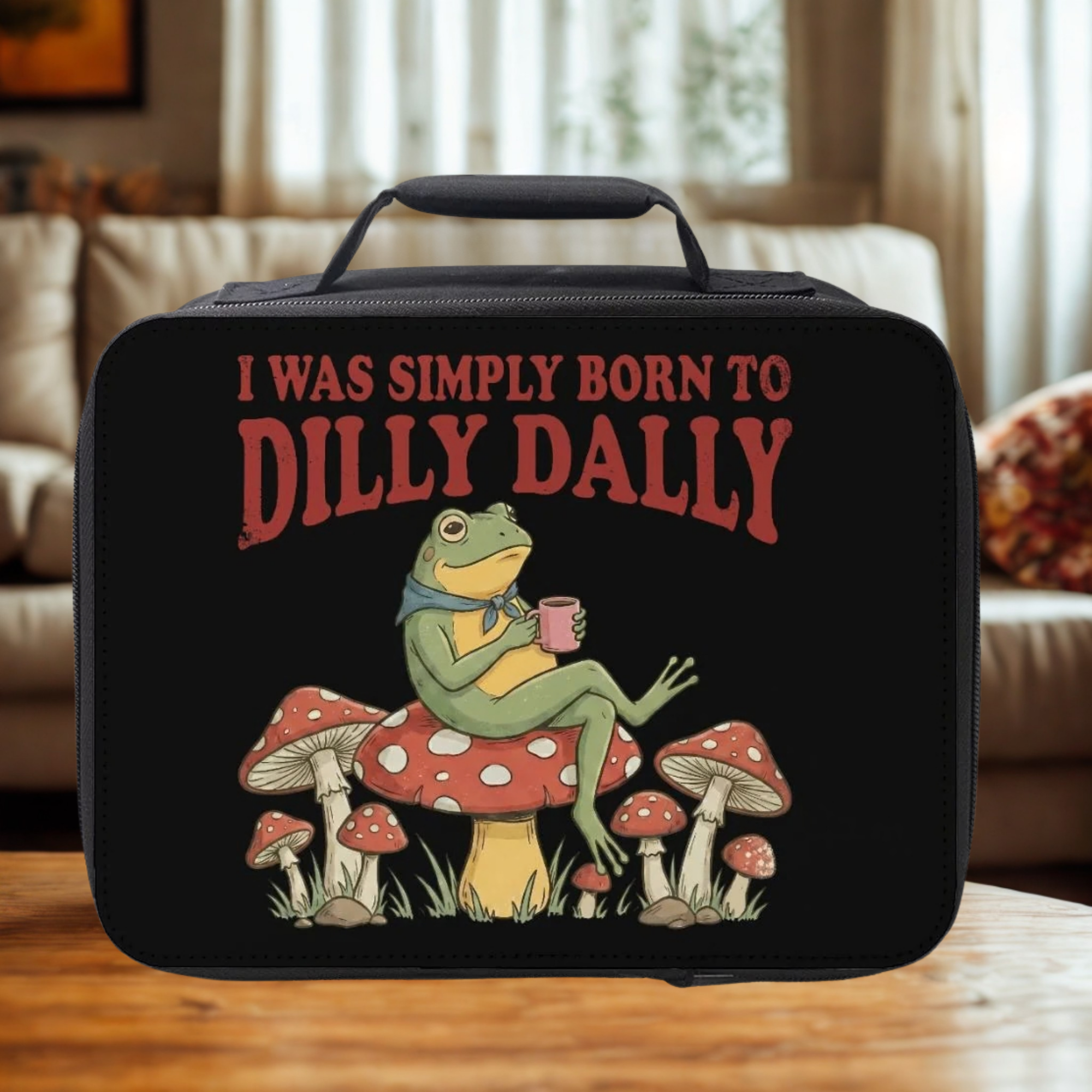 Black lunch bag with a frog graphic and text 'I was simply born to dilly dally' on a wooden living room table.