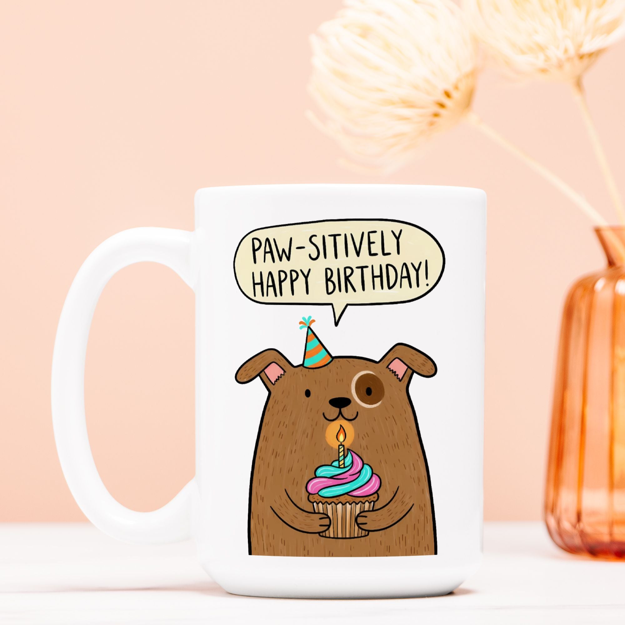 White mug with a cartoon dog holding a birthday cupcake and a speech bubble saying 'PAW-SITIVELY HAPPY BIRTHDAY!' on a light pink background.
