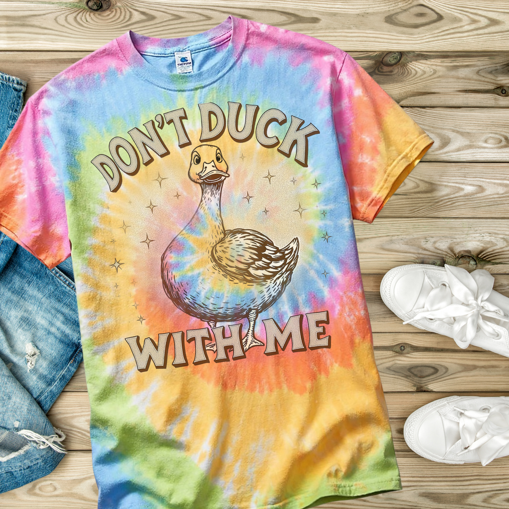 Tie-dye t-shirt with a duck graphic and text on a wooden surface with jeans and shoes.